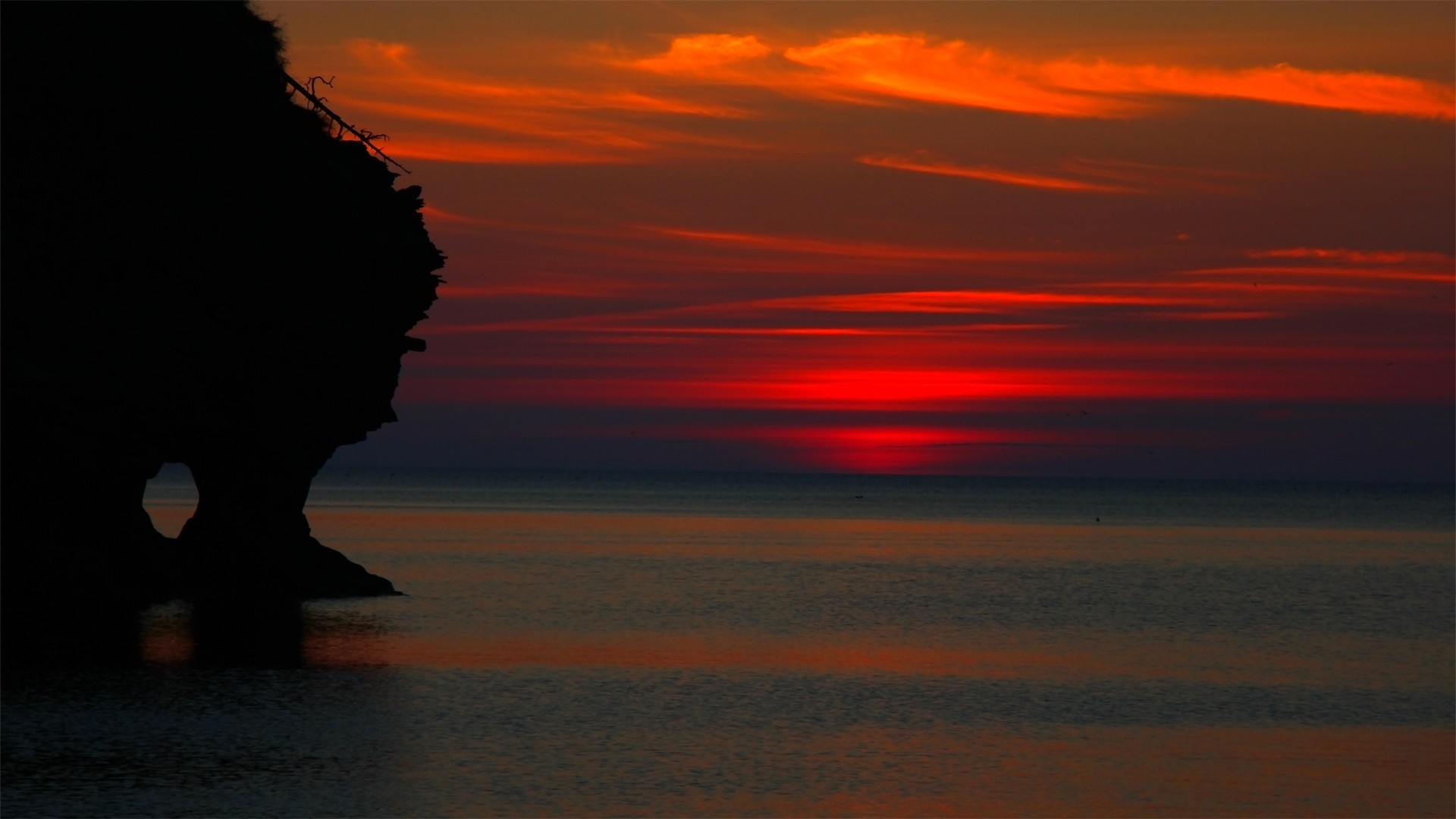 Silhouette of a rock formation against a vibrant sunset over the ocean HD Wallpaper