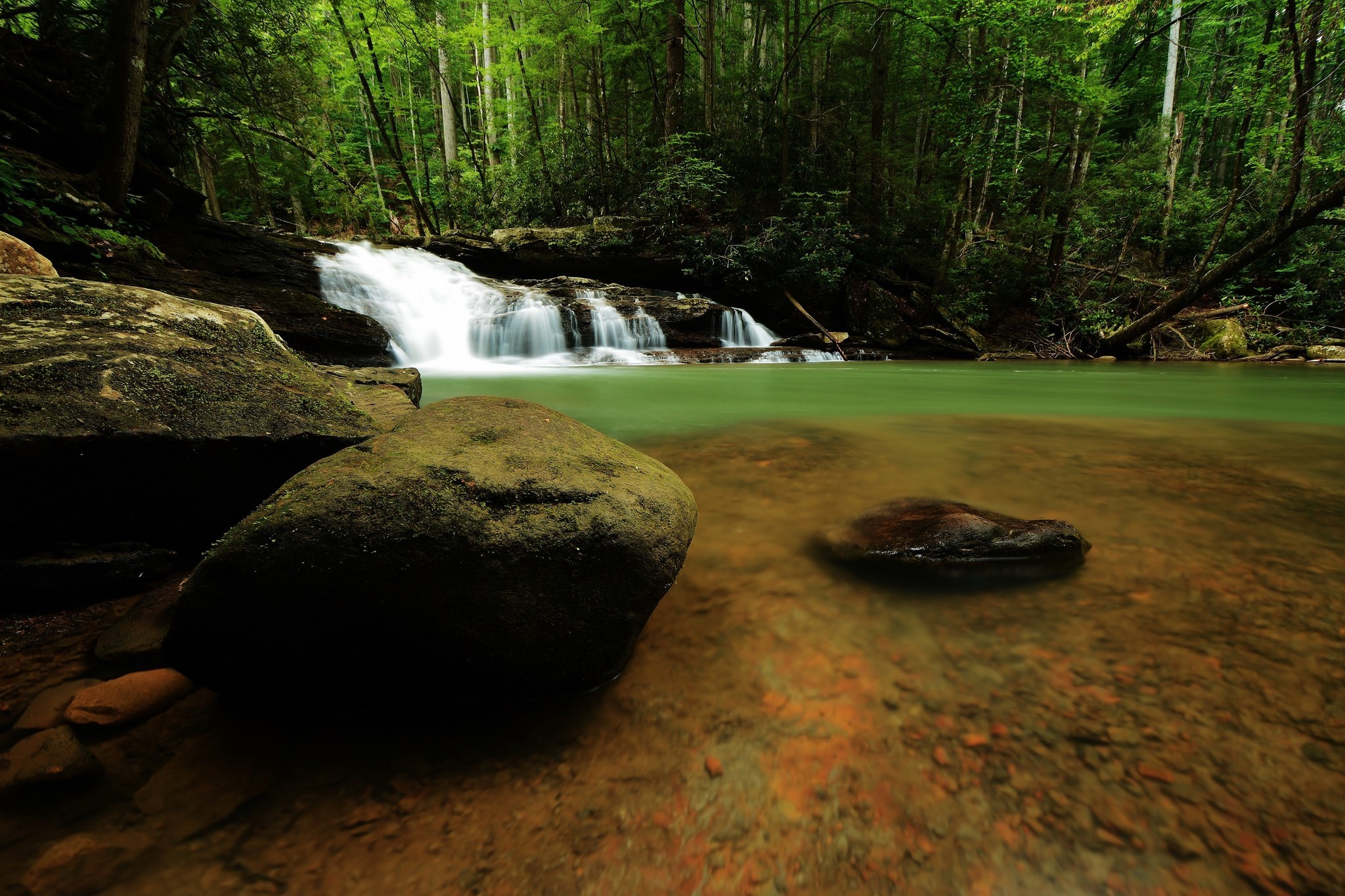 Waterfall flowing through a lush green forest with large rocks HD Wallpaper