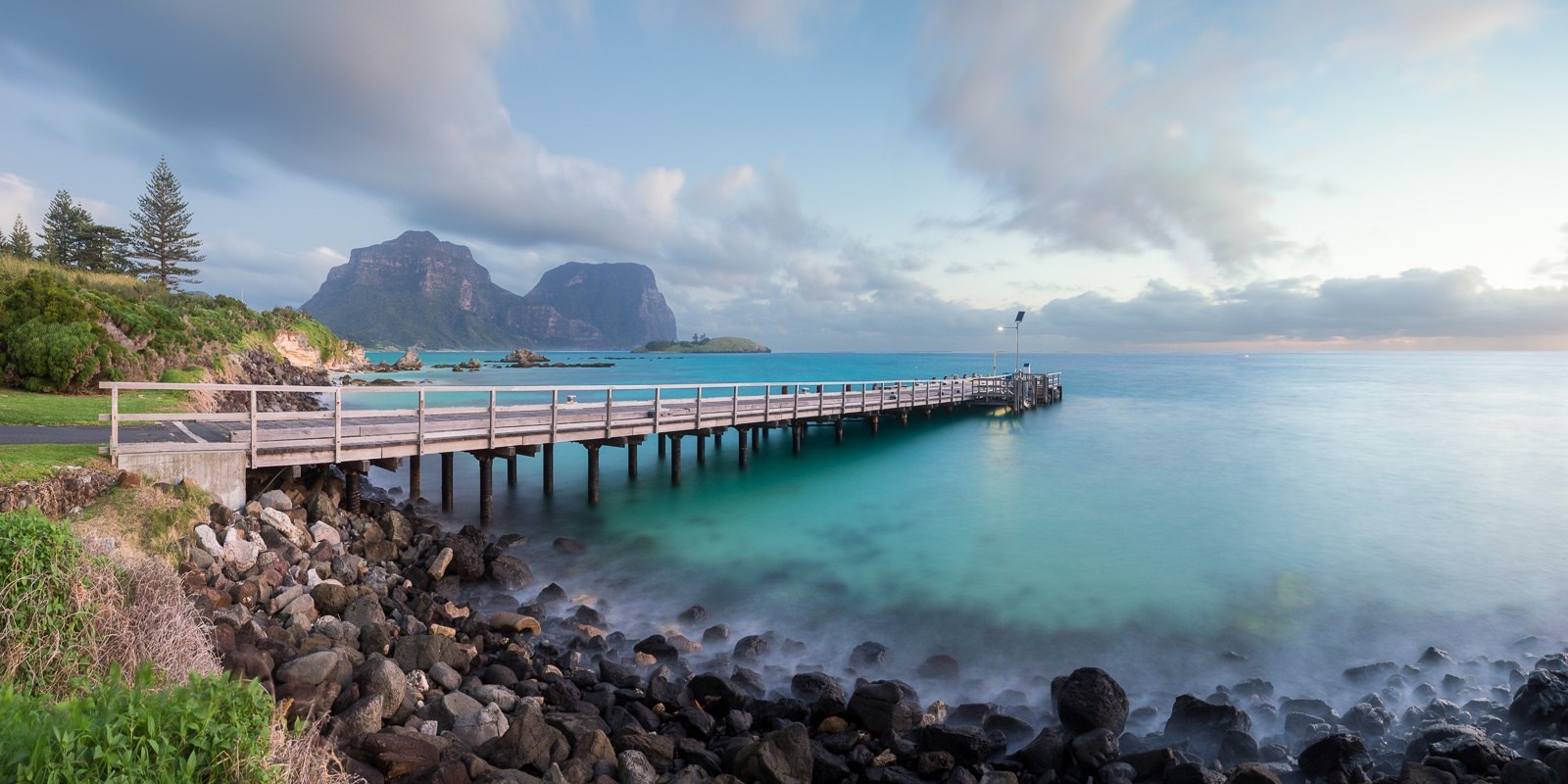 Wooden pier extending into a turquoise bay with mountains in the background HD Wallpaper