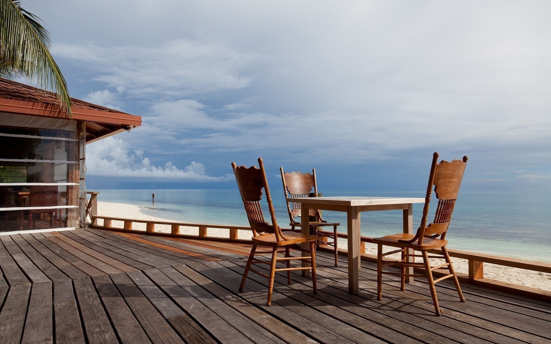 Wooden deck with table and chairs overlooking a tropical beach and ocean HD Wallpaper