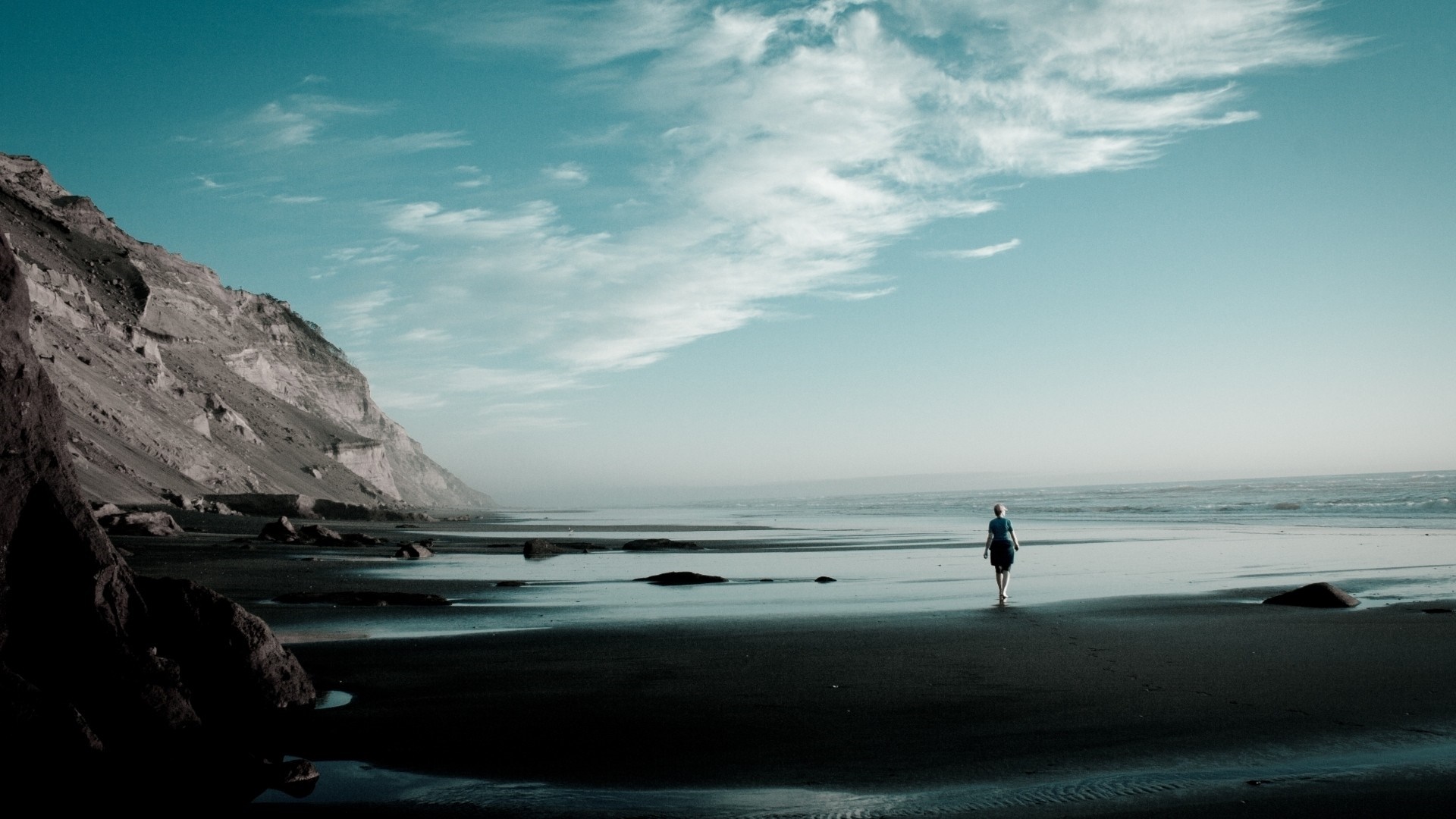Woman walking on a dark beach with cliffs and ocean HD Wallpaper