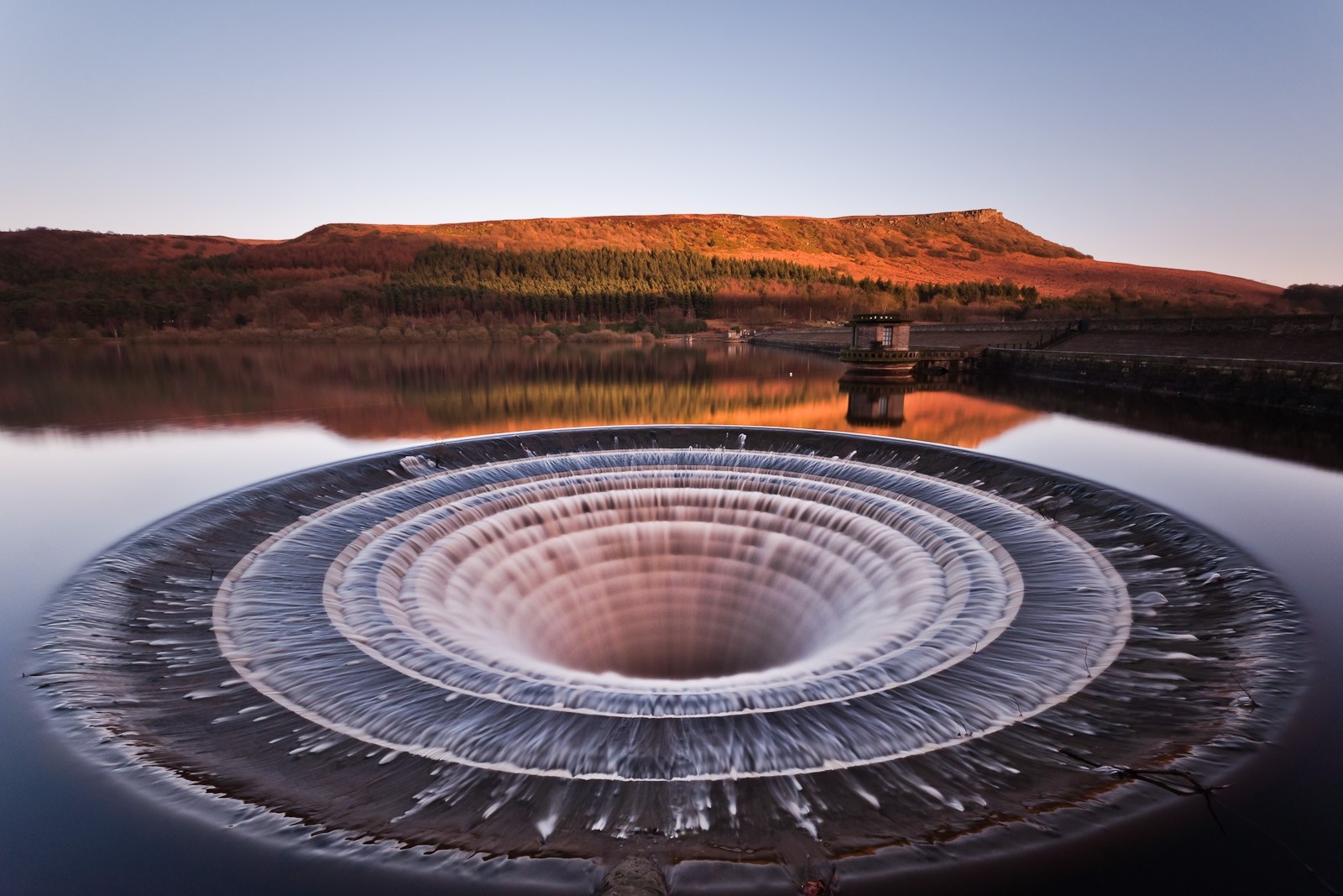 Circular overflow drain in a reservoir with hills in the background HD Wallpaper
