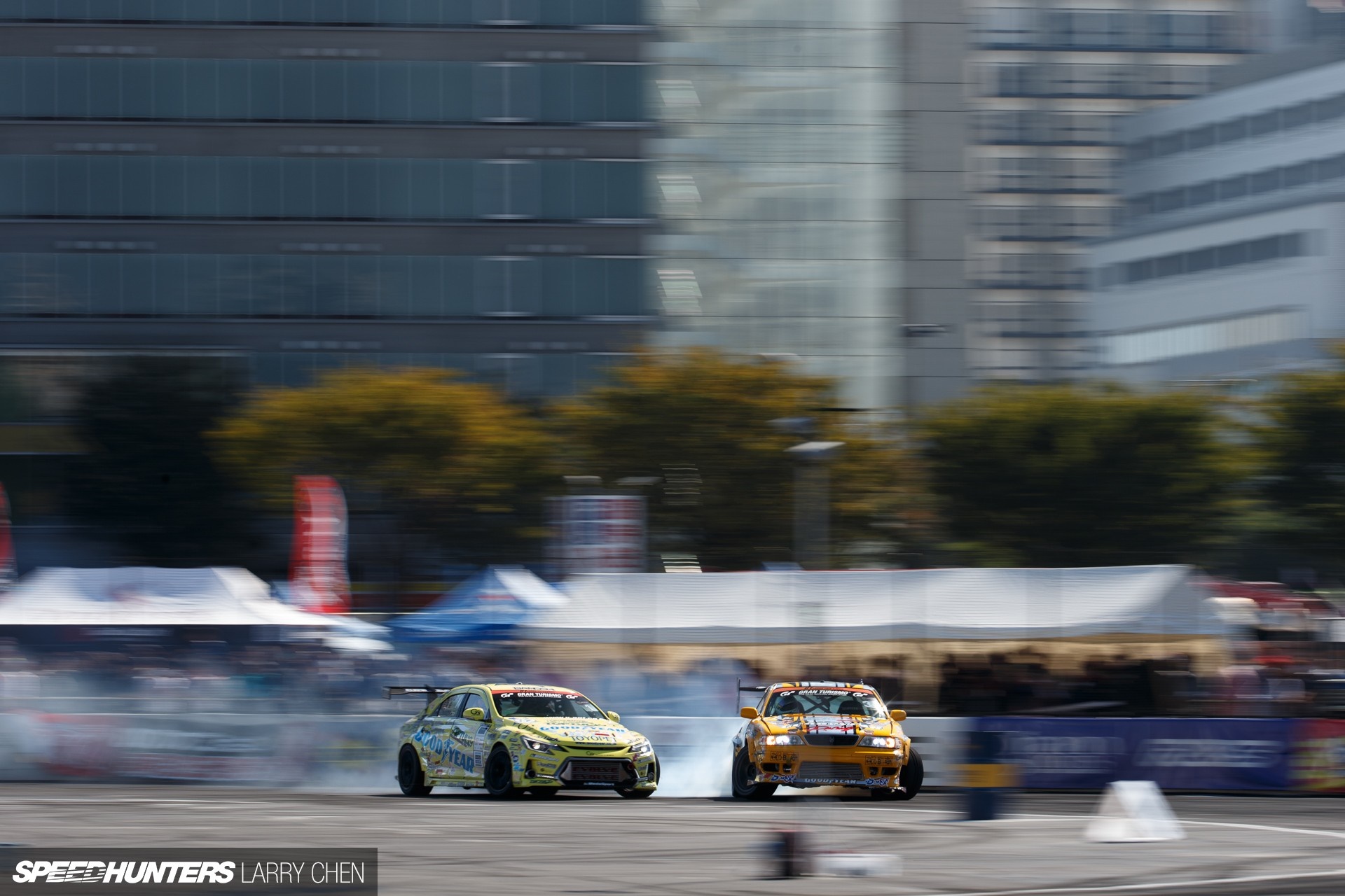 Two drift cars racing on a track with buildings in background HD Wallpaper