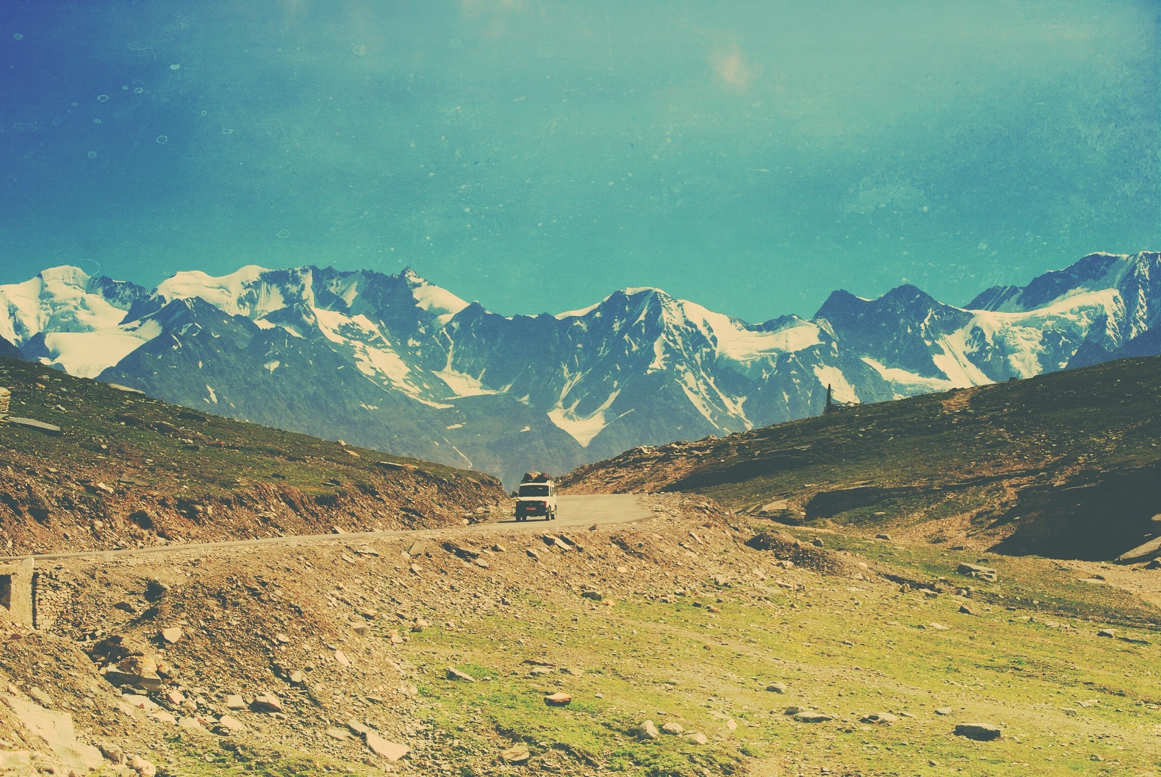 A car drives on a mountain road with snow-capped mountains in the background. HD Wallpaper