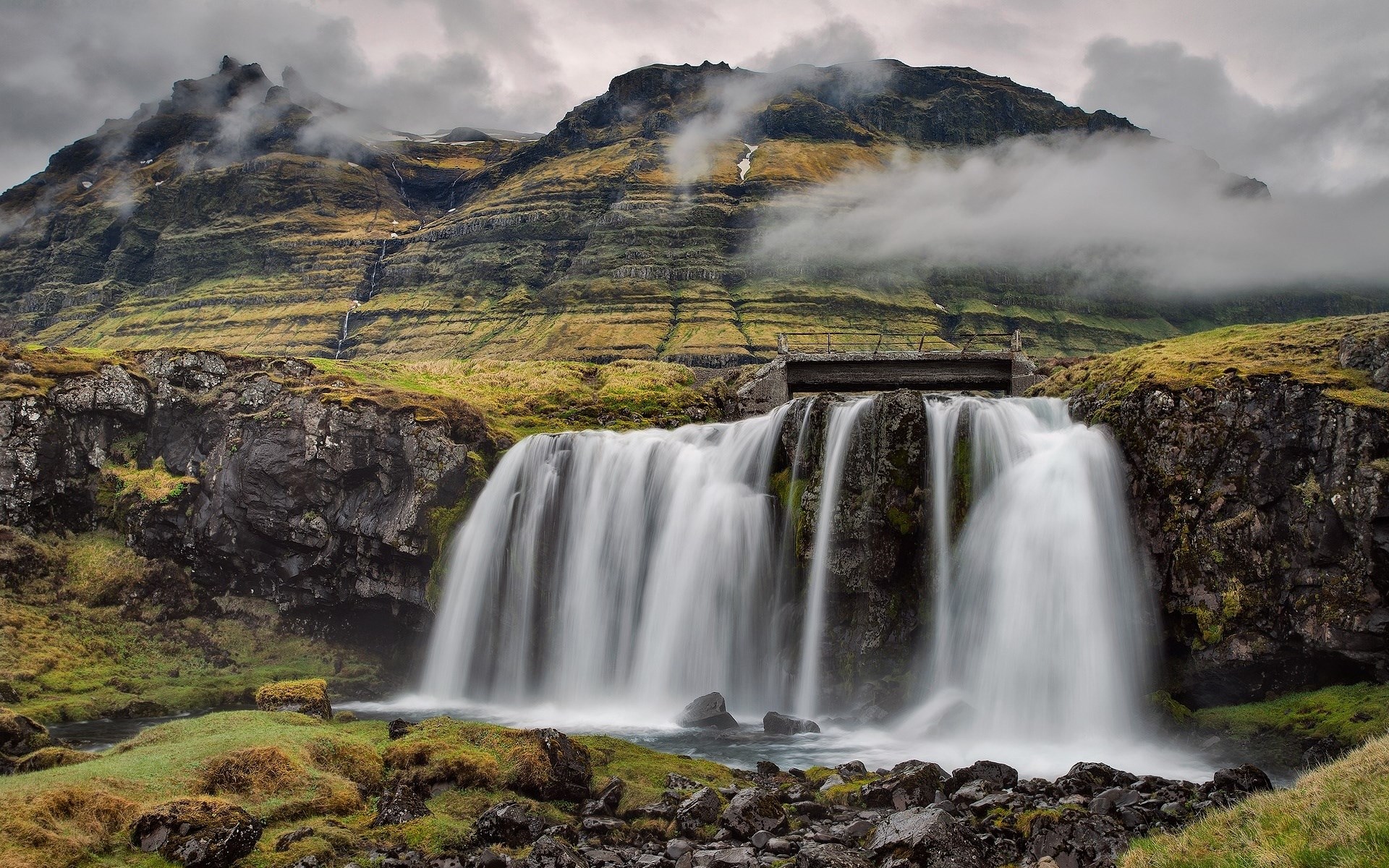 Waterfall cascading down a rocky cliff face with mountains in the background HD Wallpaper