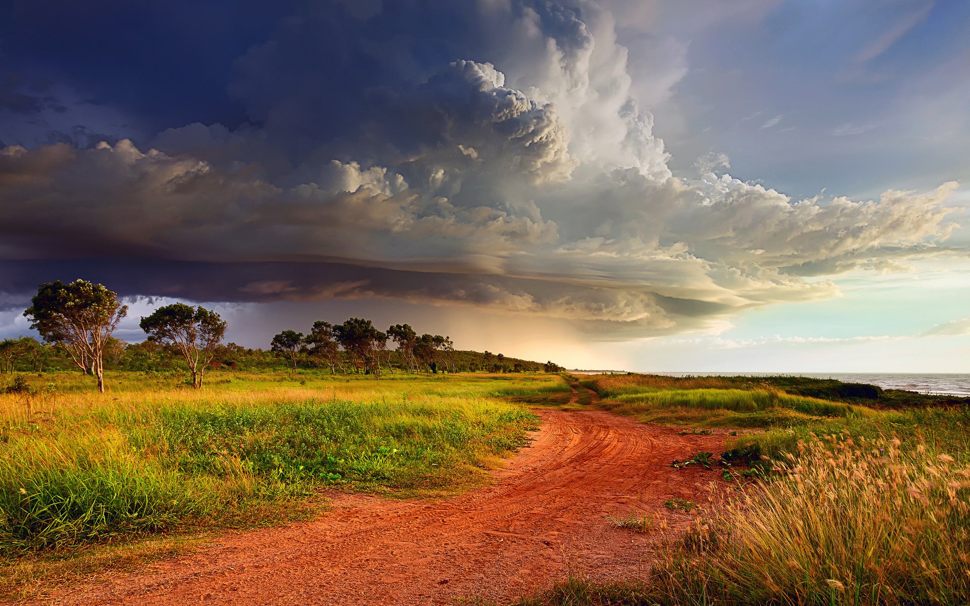 Dirt road through grassy field towards the sea under stormy clouds HD Wallpaper