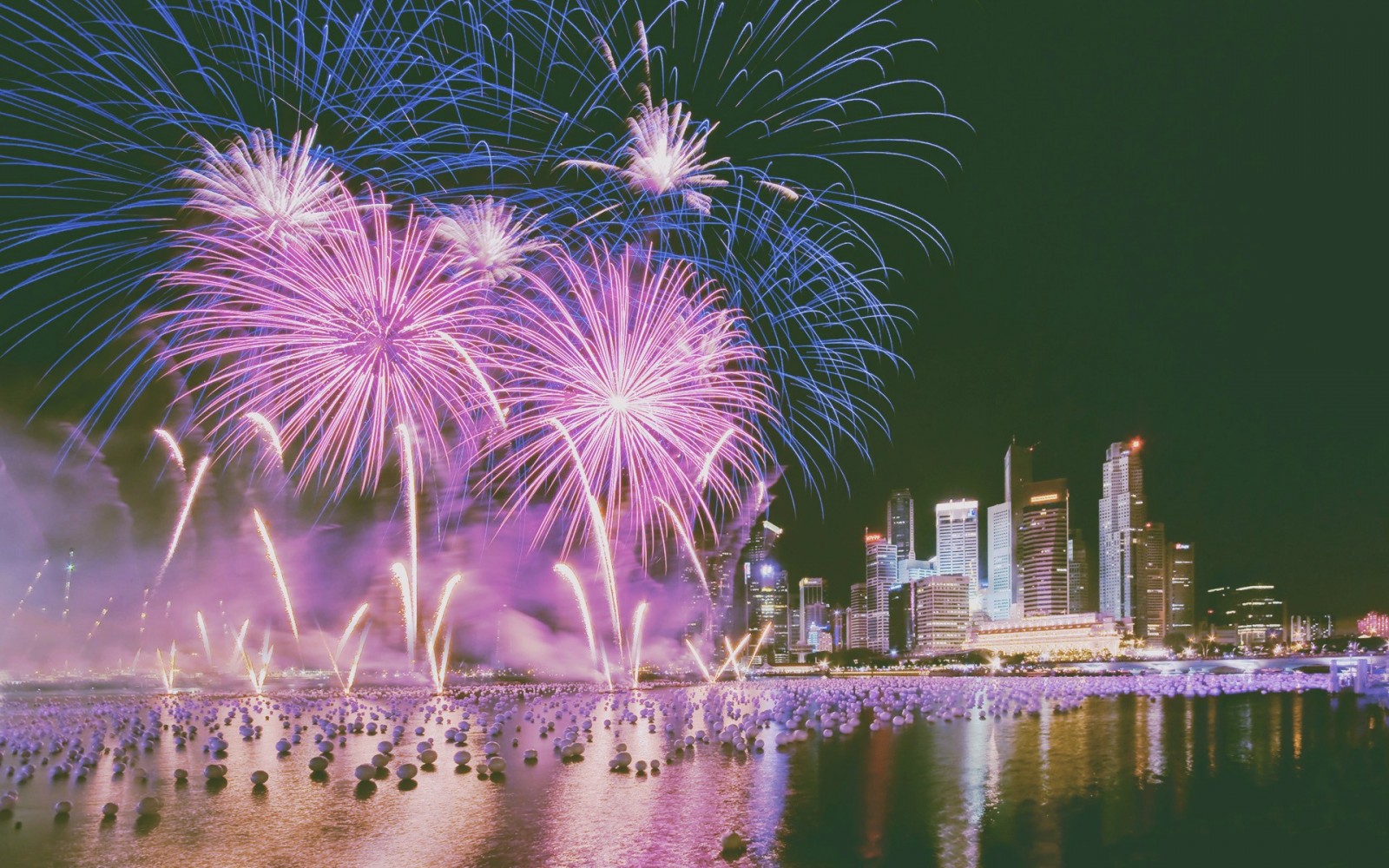 Fireworks display over a city skyline and water HD Wallpaper