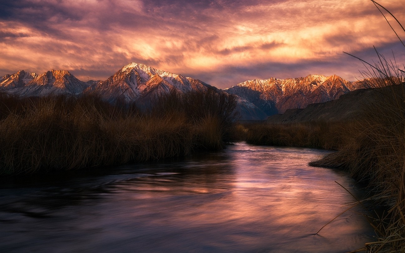 River flowing through marshland with snow-capped mountains at sunset HD Wallpaper