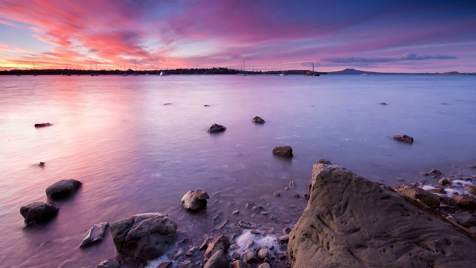 Sunset over water with rocks and distant land HD Wallpaper