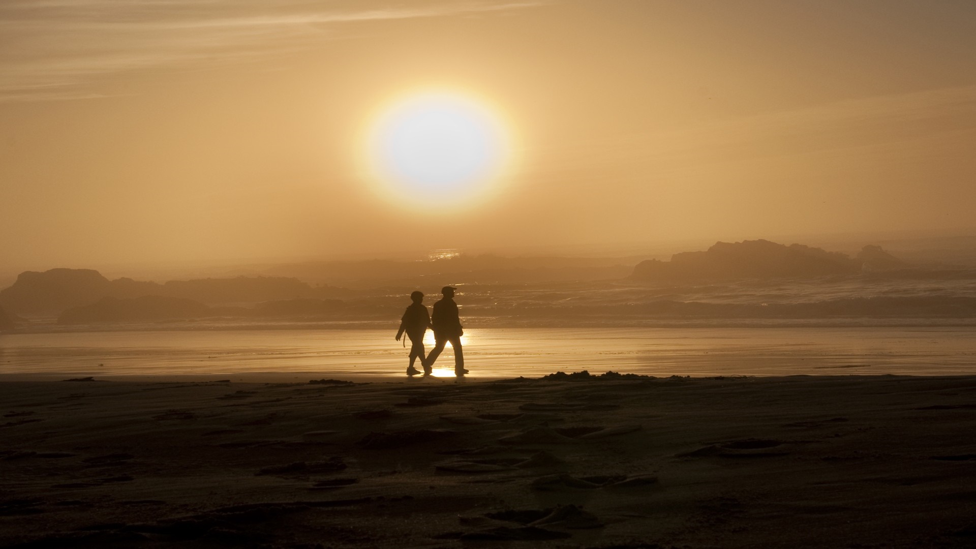 Two people walking on a beach at sunset HD Wallpaper