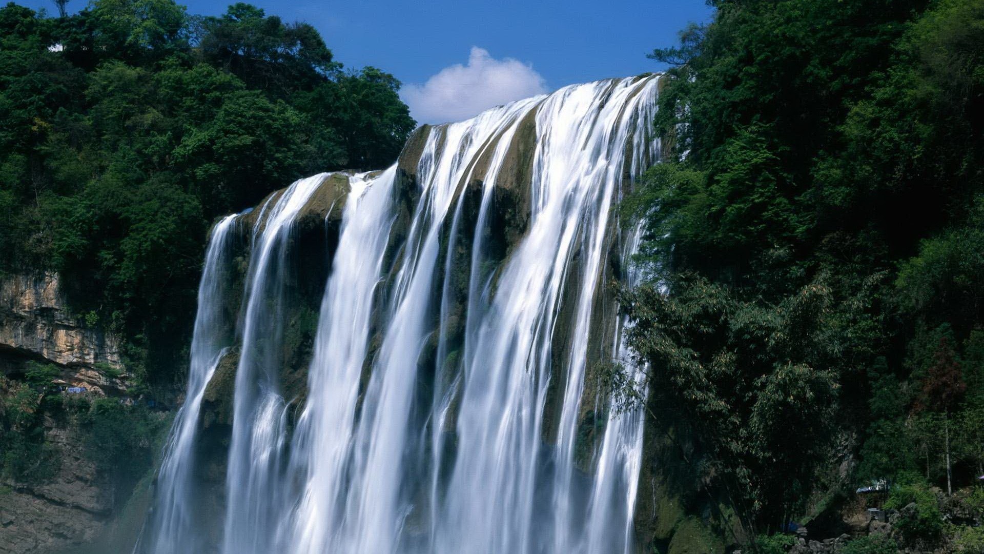 A wide waterfall cascades down a rocky cliff surrounded by trees HD Wallpaper