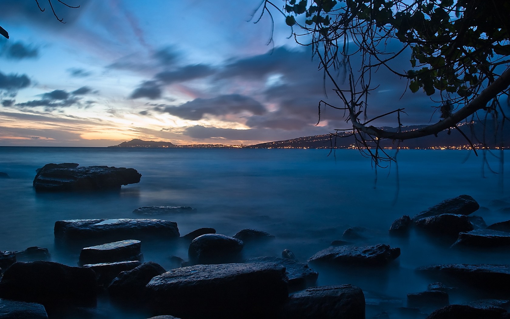 Rocks in the ocean at dusk with city lights on the horizon HD Wallpaper