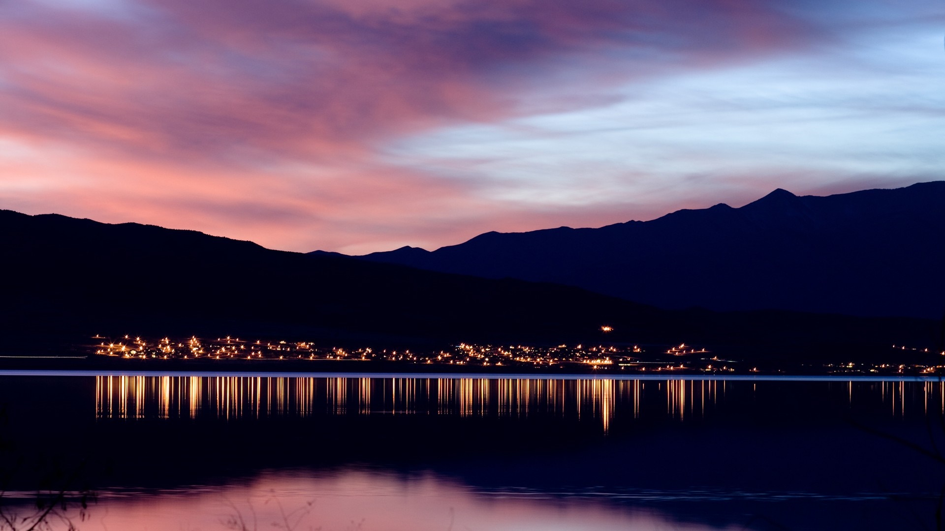 City lights reflected on a lake at sunset with mountains HD Wallpaper