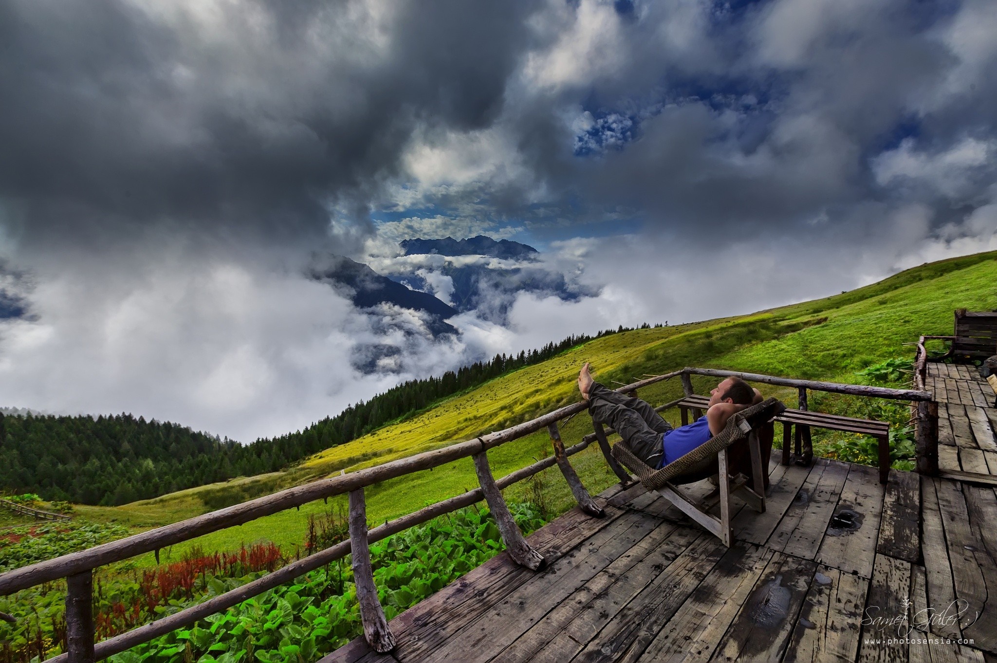 Person relaxing on a wooden deck overlooking a cloudy mountain valley HD Wallpaper