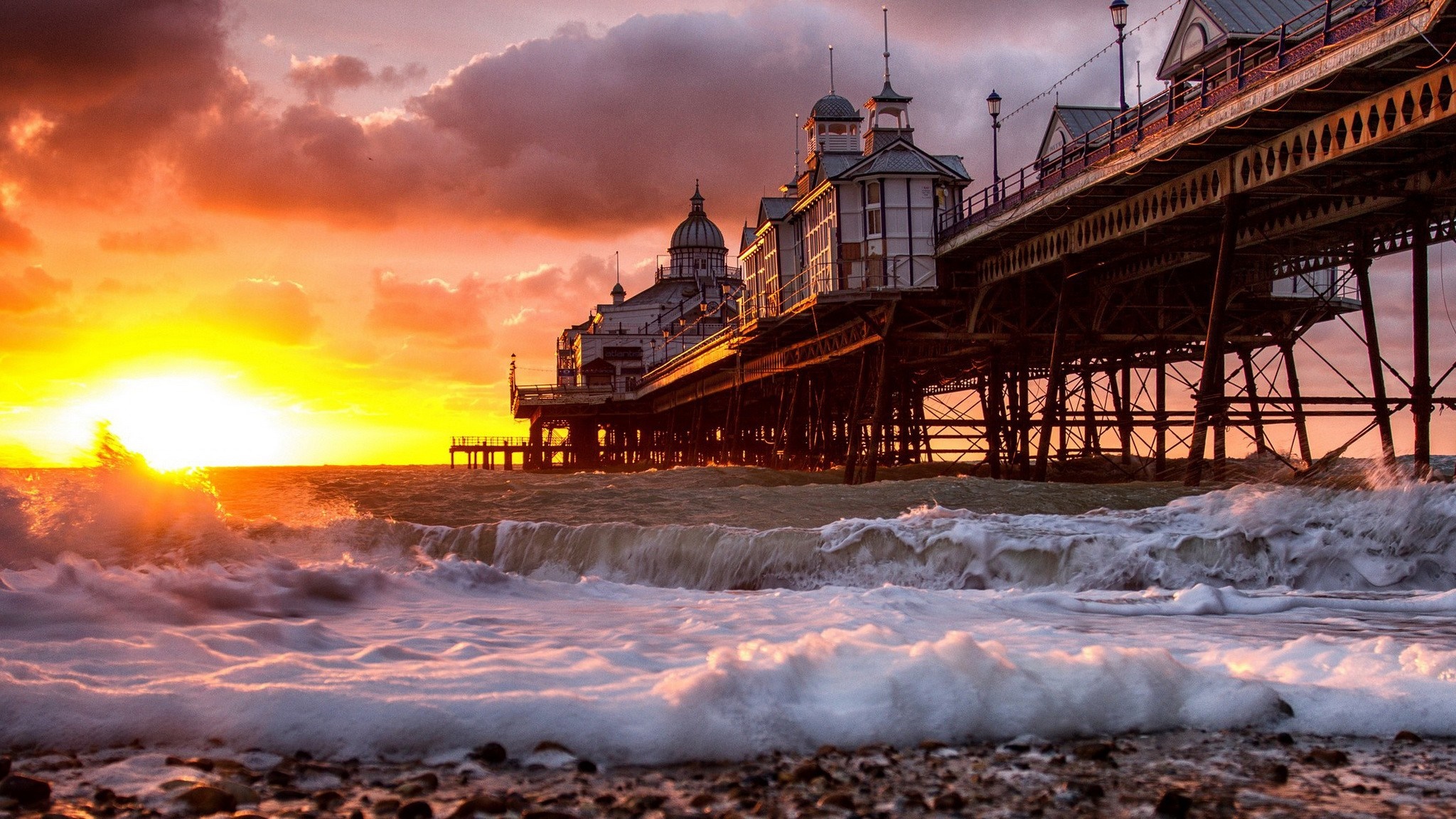 Sunset over a pier with waves crashing on the shore HD Wallpaper