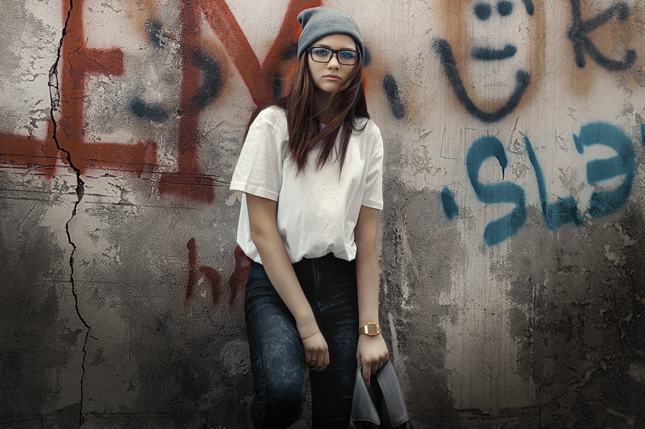Young woman in glasses and beanie leaning against graffiti wall HD Wallpaper