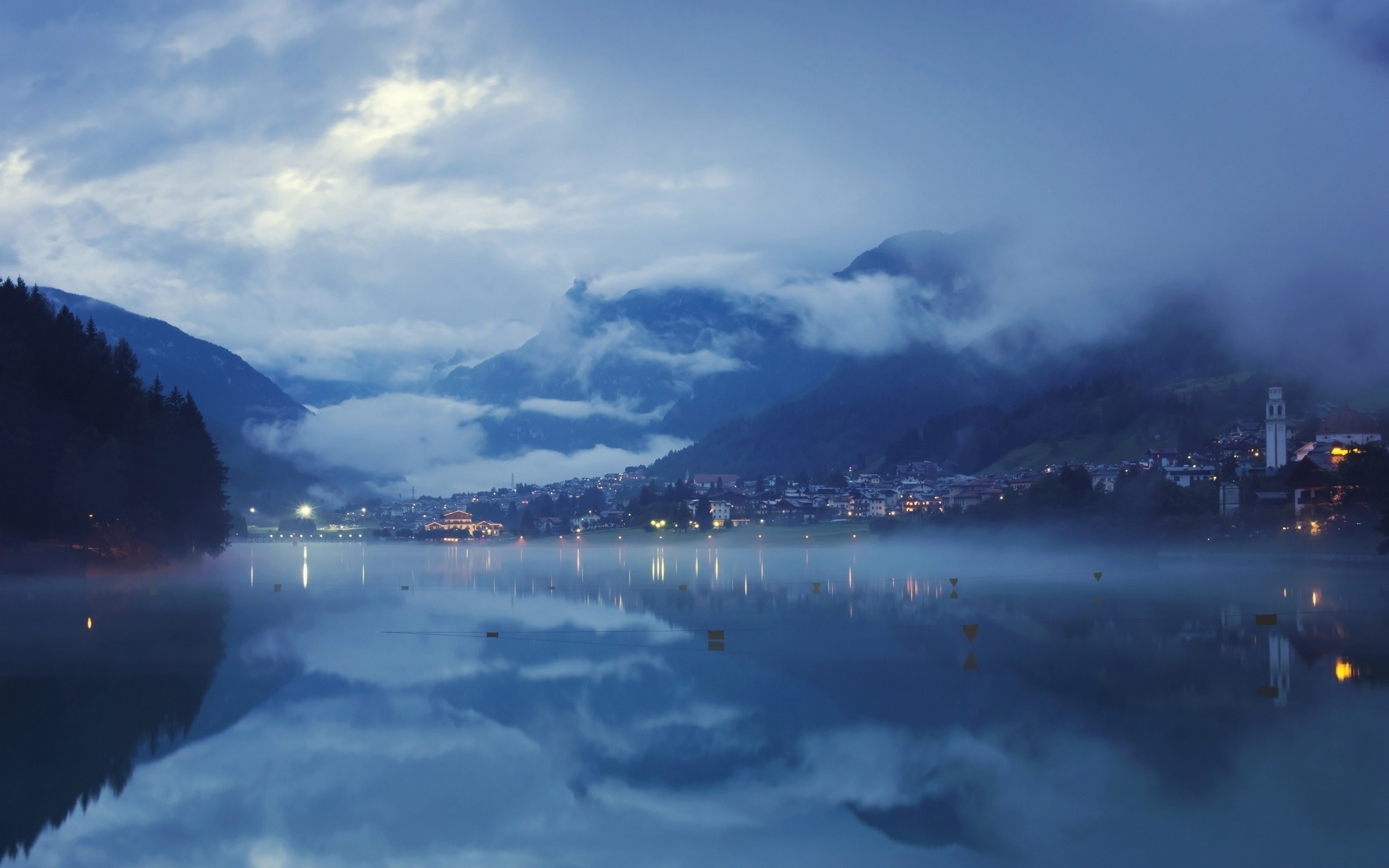 Misty lake reflecting mountains and a town at dusk HD Wallpaper