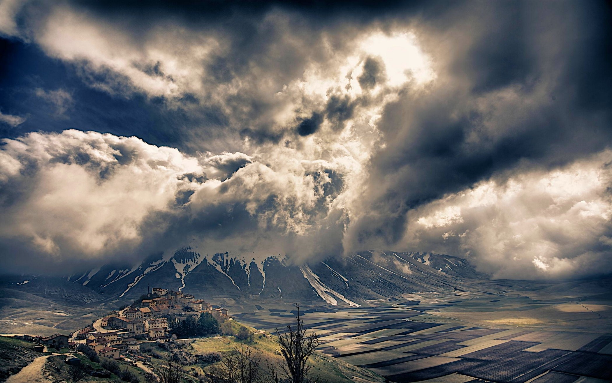 Village on a hill under stormy clouds and snowy mountains HD Wallpaper
