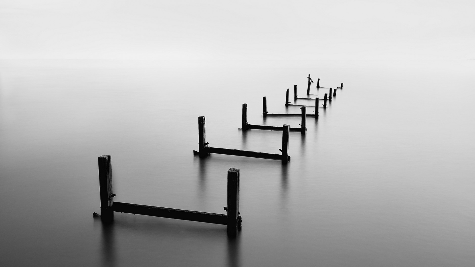 Monochrome long exposure of wooden pier remnants in calm water HD Wallpaper