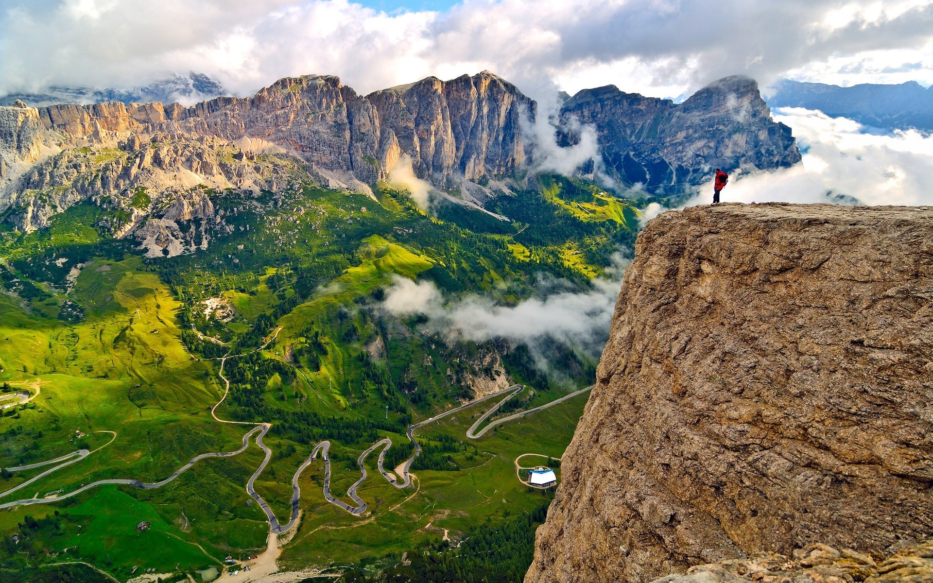 Person on cliff overlooking winding mountain road in Alps HD Wallpaper