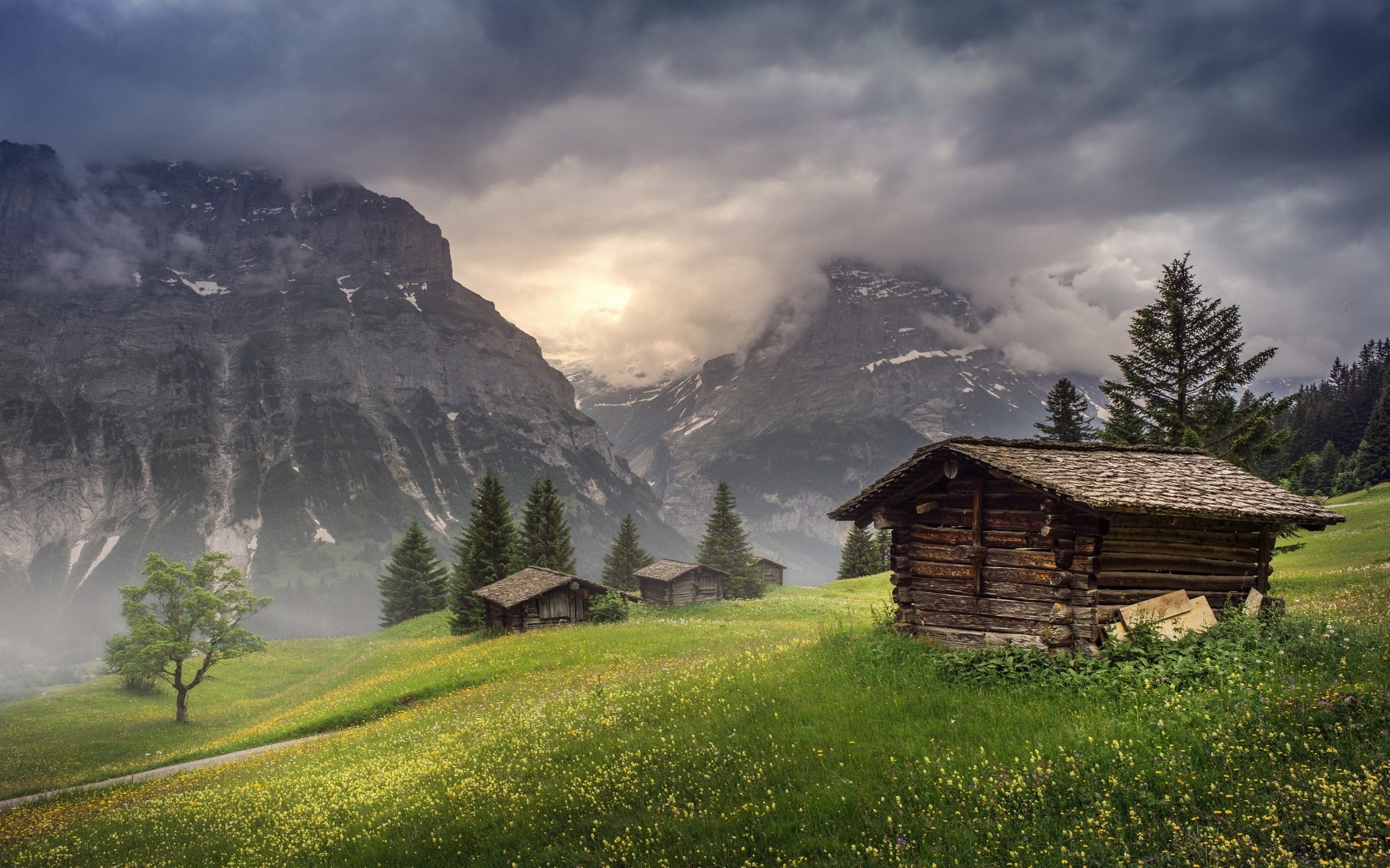 Wooden huts in a green meadow with mountains and clouds HD Wallpaper