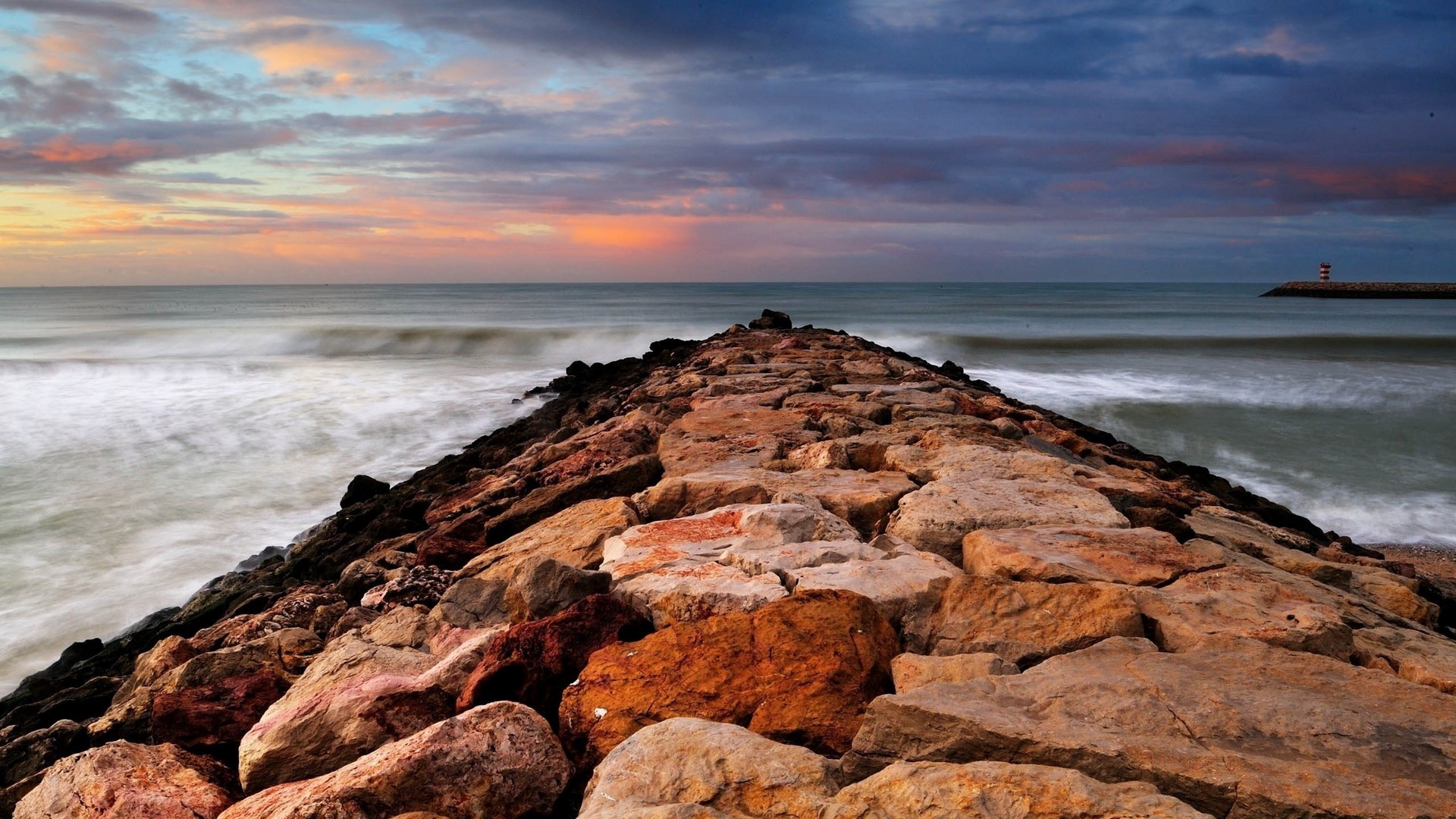 Stone jetty leading to a lighthouse at sunset HD Wallpaper