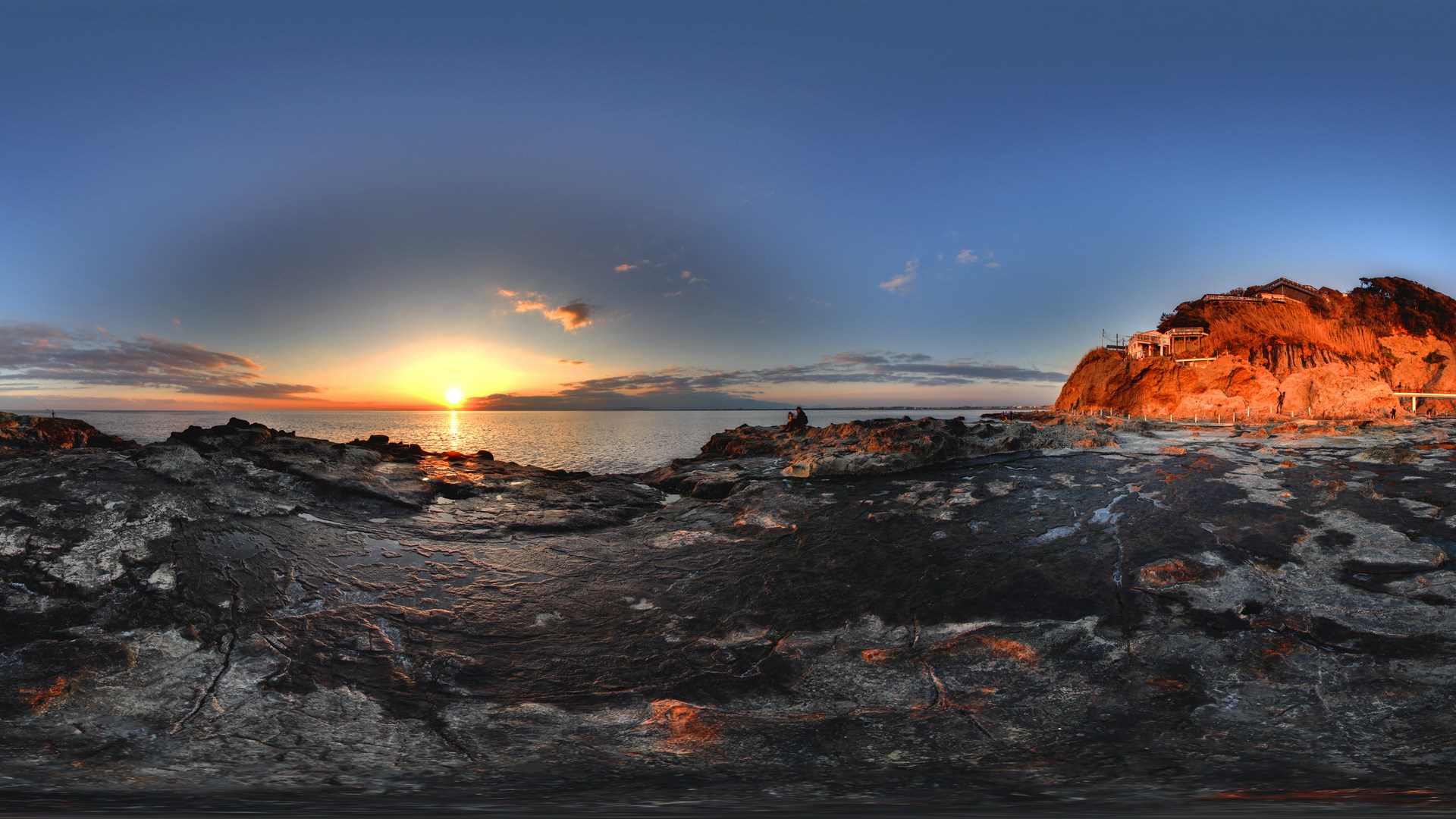 Sunset over the ocean with rocky shore and cliffside buildings HD Wallpaper