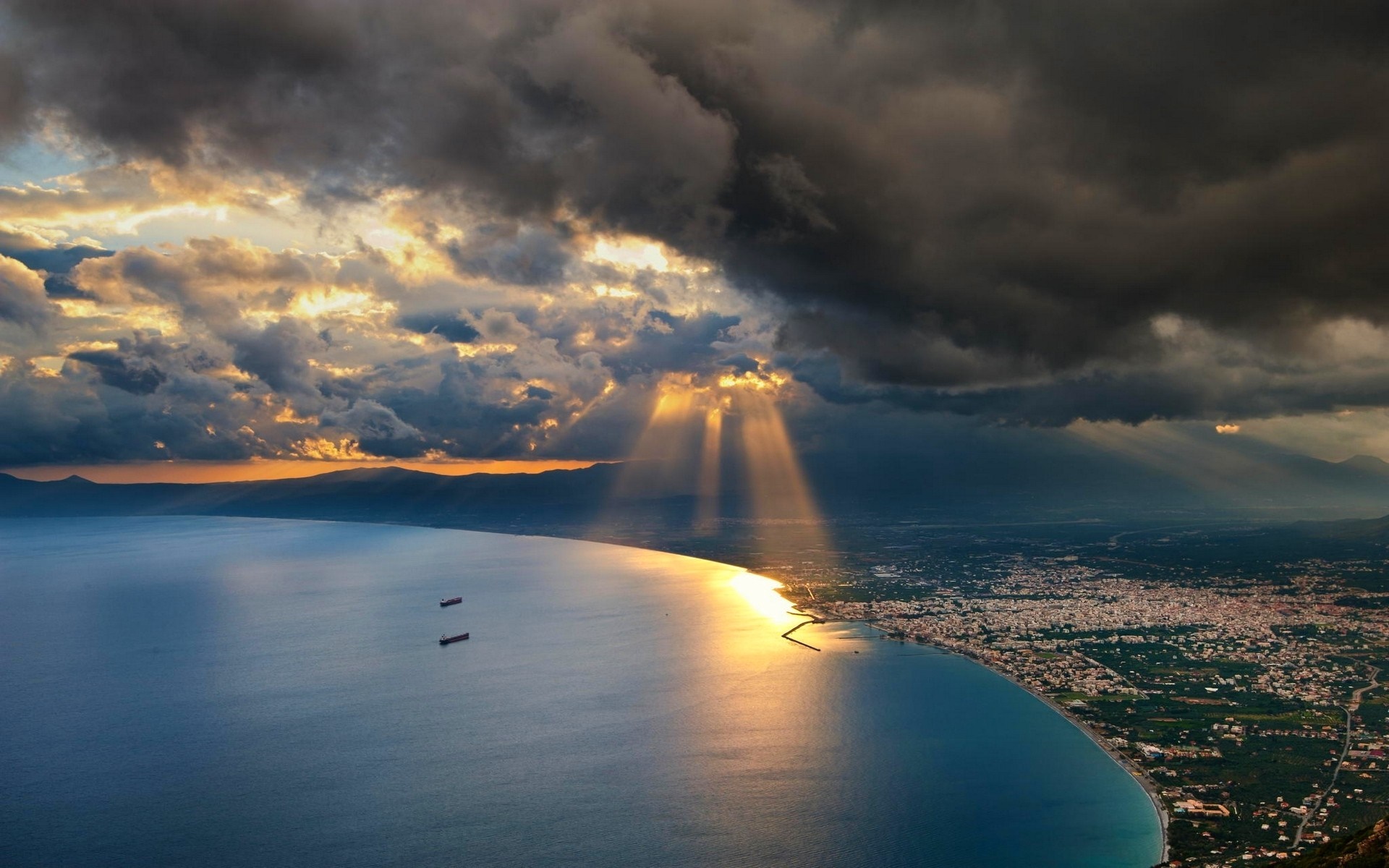 Sun rays breaking through clouds over a coastal cityscape and sea HD Wallpaper