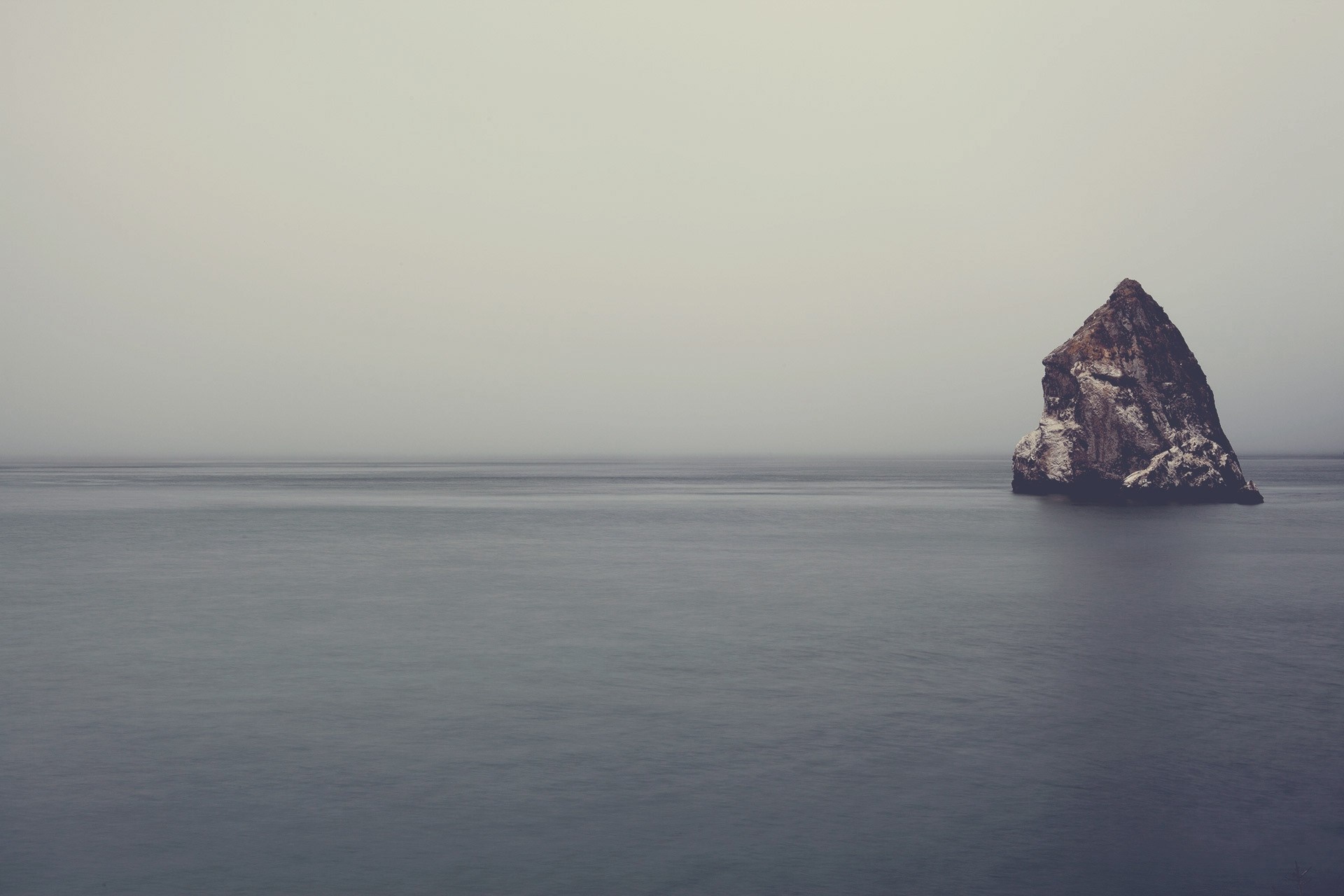 A solitary rock formation in the sea under a foggy sky HD Wallpaper