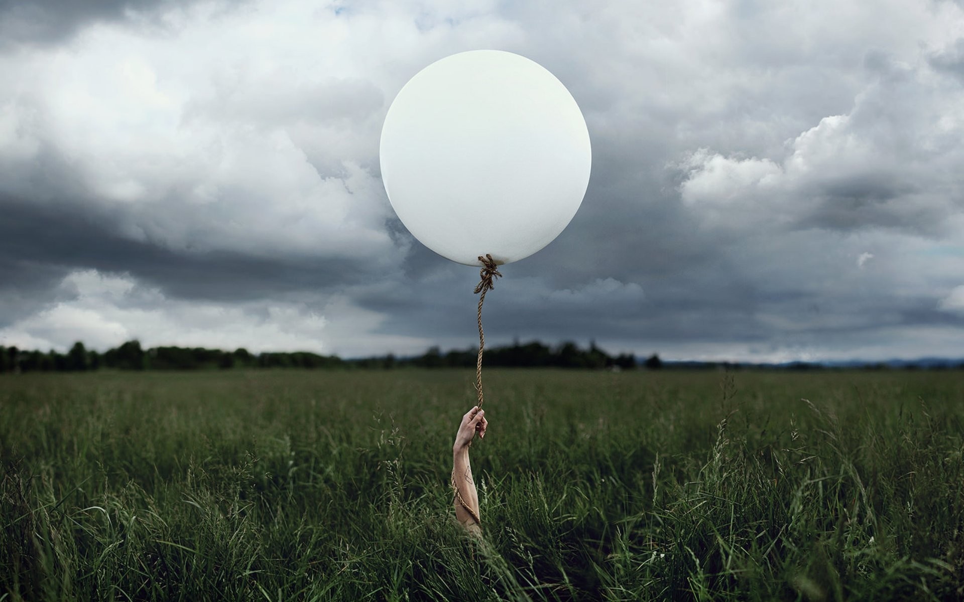 Hand holding a white balloon in a grassy field under stormy clouds HD Wallpaper