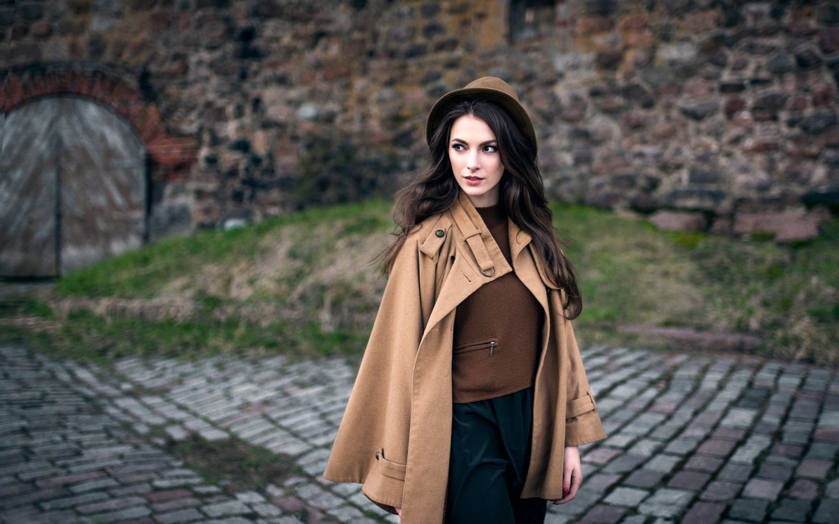 Woman in a brown coat and hat in front of a stone wall HD Wallpaper
