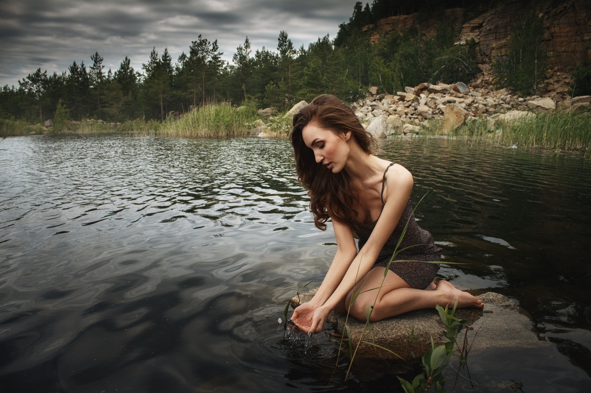 Woman kneeling by a lake with trees and rocks HD Wallpaper