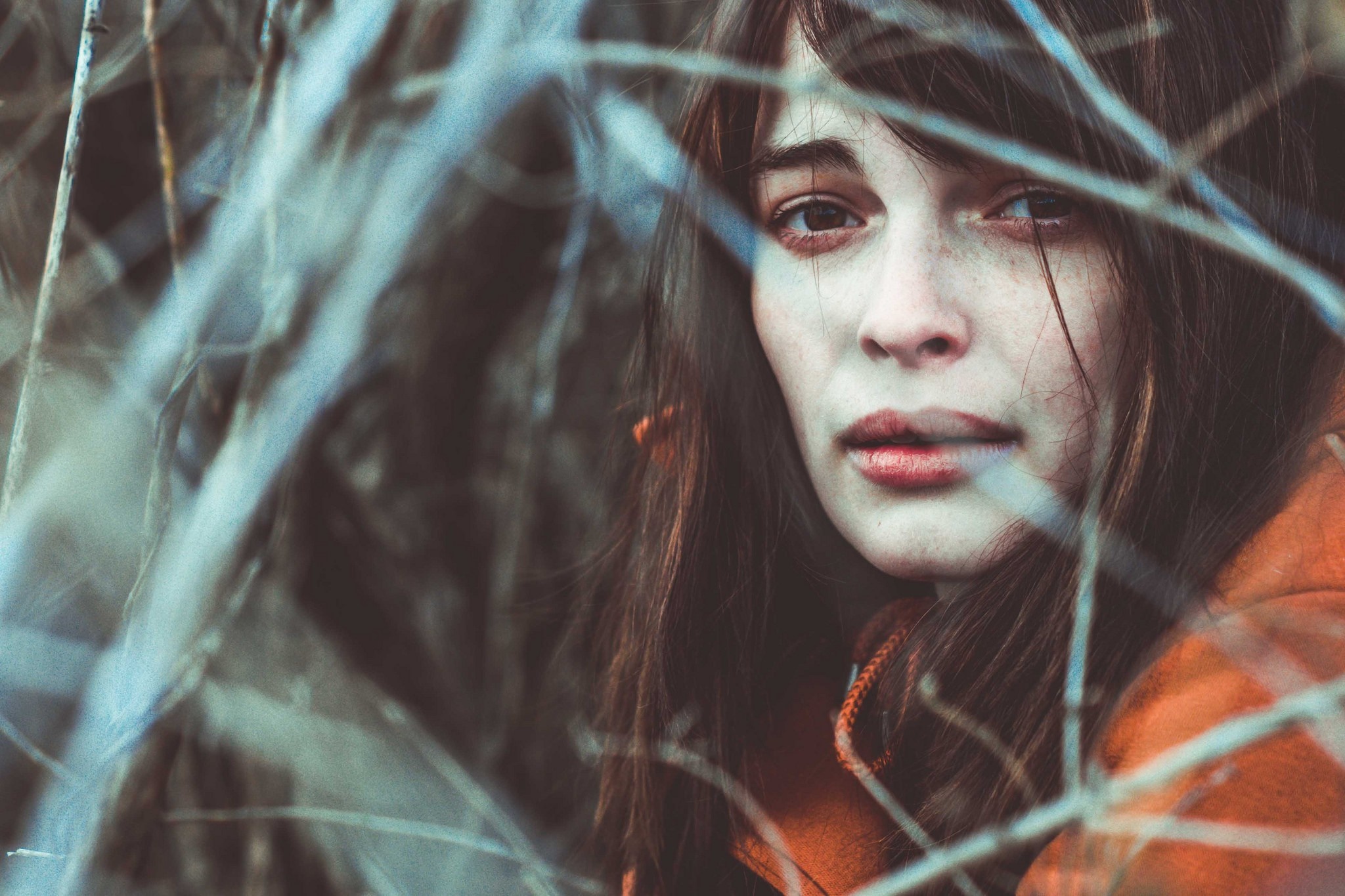 Woman with brown hair and orange jacket behind branches HD Wallpaper
