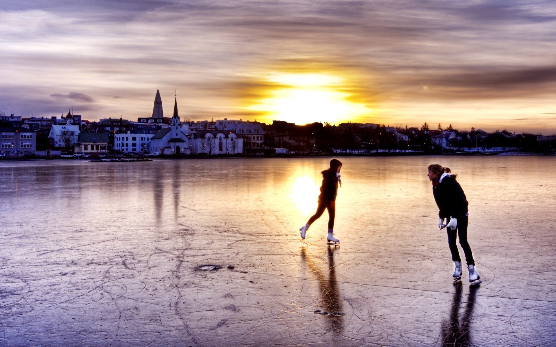 Two people ice skating on a frozen lake at sunset with a cityscape in the background. HD Wallpaper
