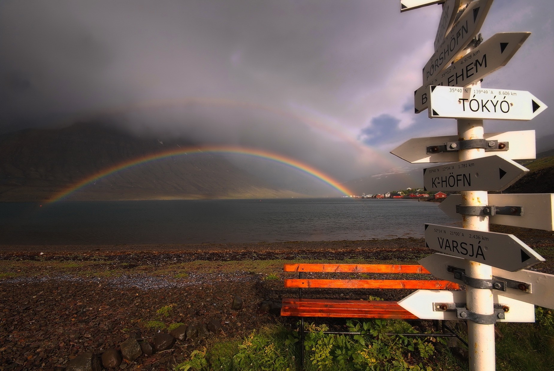 Rainbow over a bay with a signpost and bench HD Wallpaper