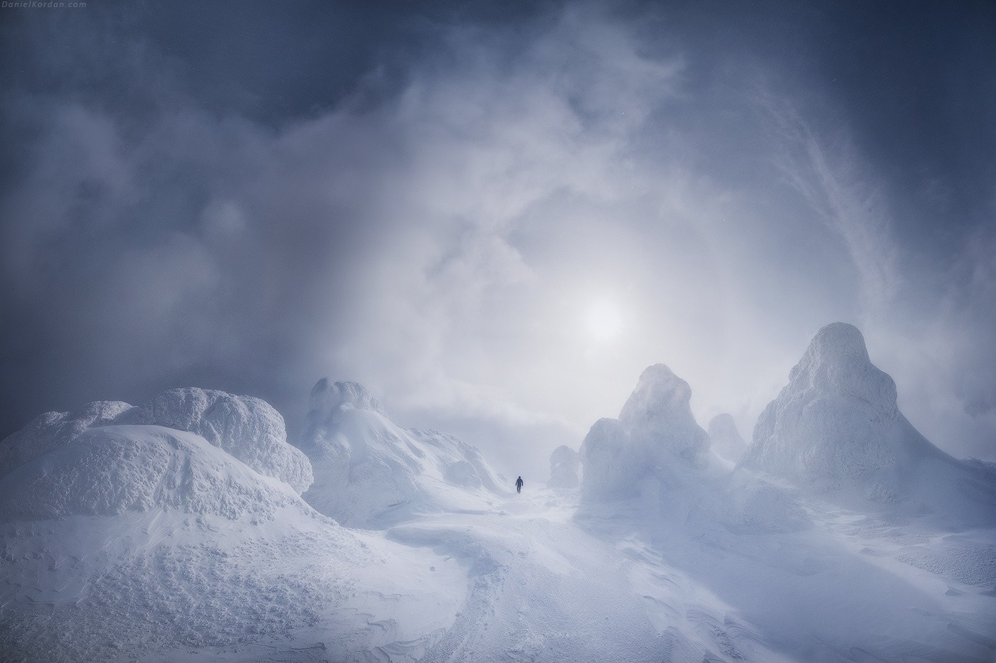 Person walking through a snowy, icy mountain landscape under a cloudy sky HD Wallpaper