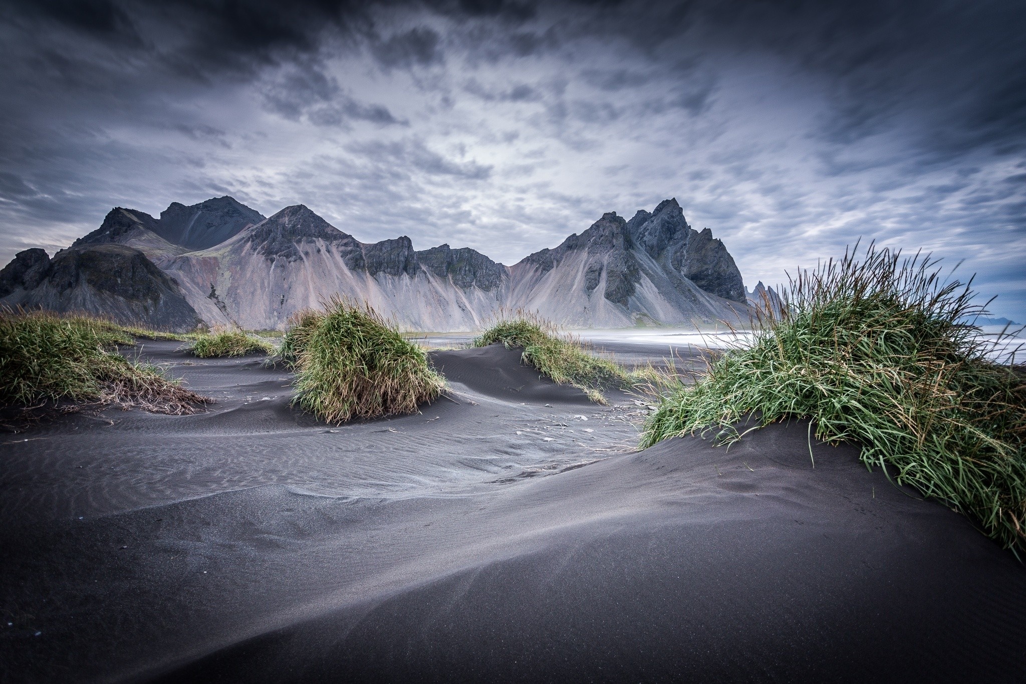 Black sand beach with mountains and grass under cloudy sky HD Wallpaper