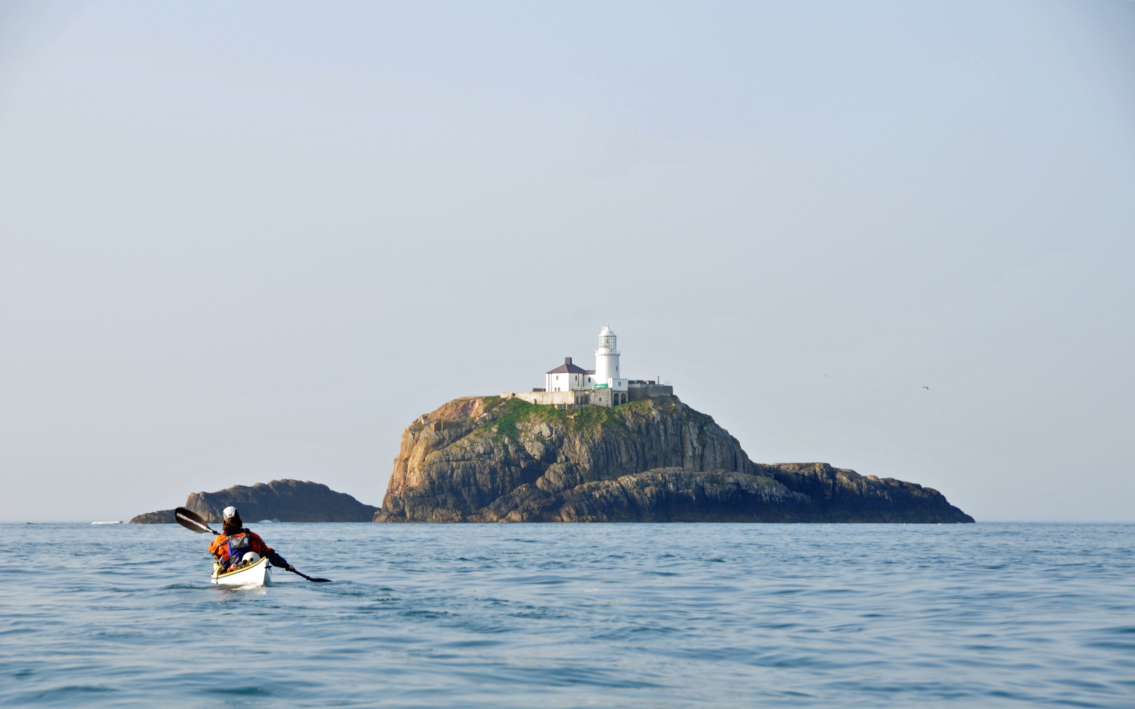 Kayaker paddles towards a lighthouse on a rocky island HD Wallpaper