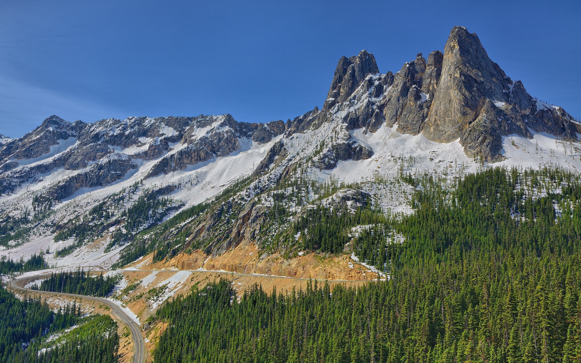 Mountain road winding through snow-capped peaks and pine forest HD Wallpaper