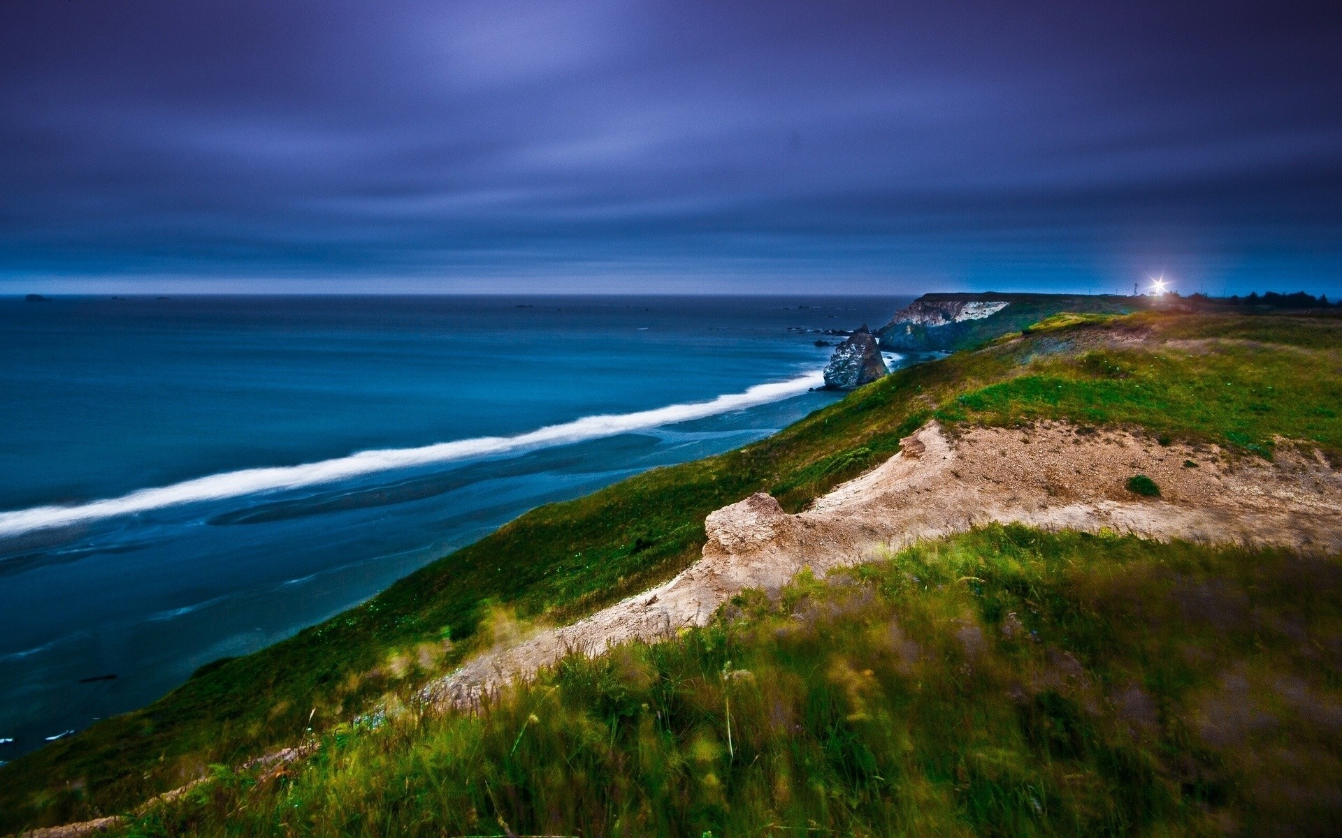 Coastal cliff overlooking the ocean at dusk with a lighthouse HD Wallpaper
