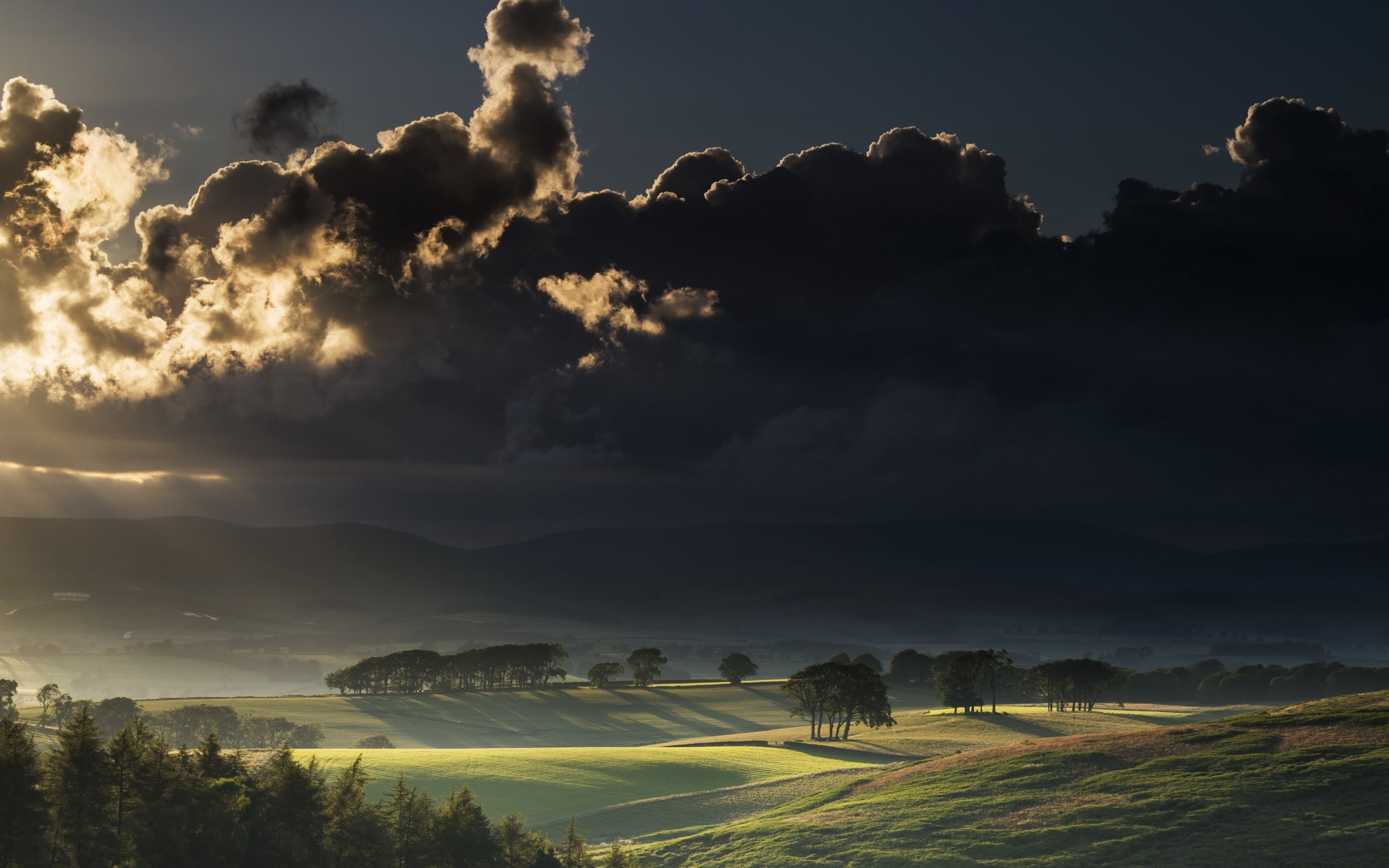 Sunlight breaking through dark clouds over rolling hills HD Wallpaper