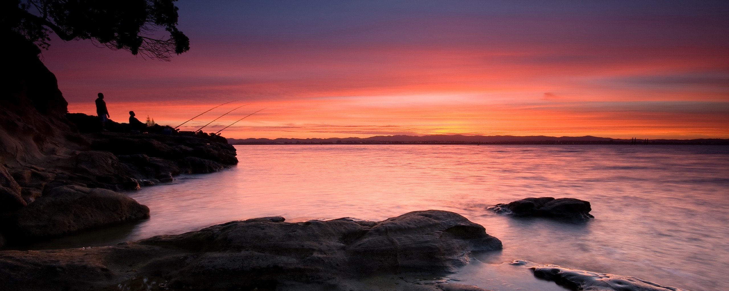 Two people fishing on a rocky shore at sunset HD Wallpaper