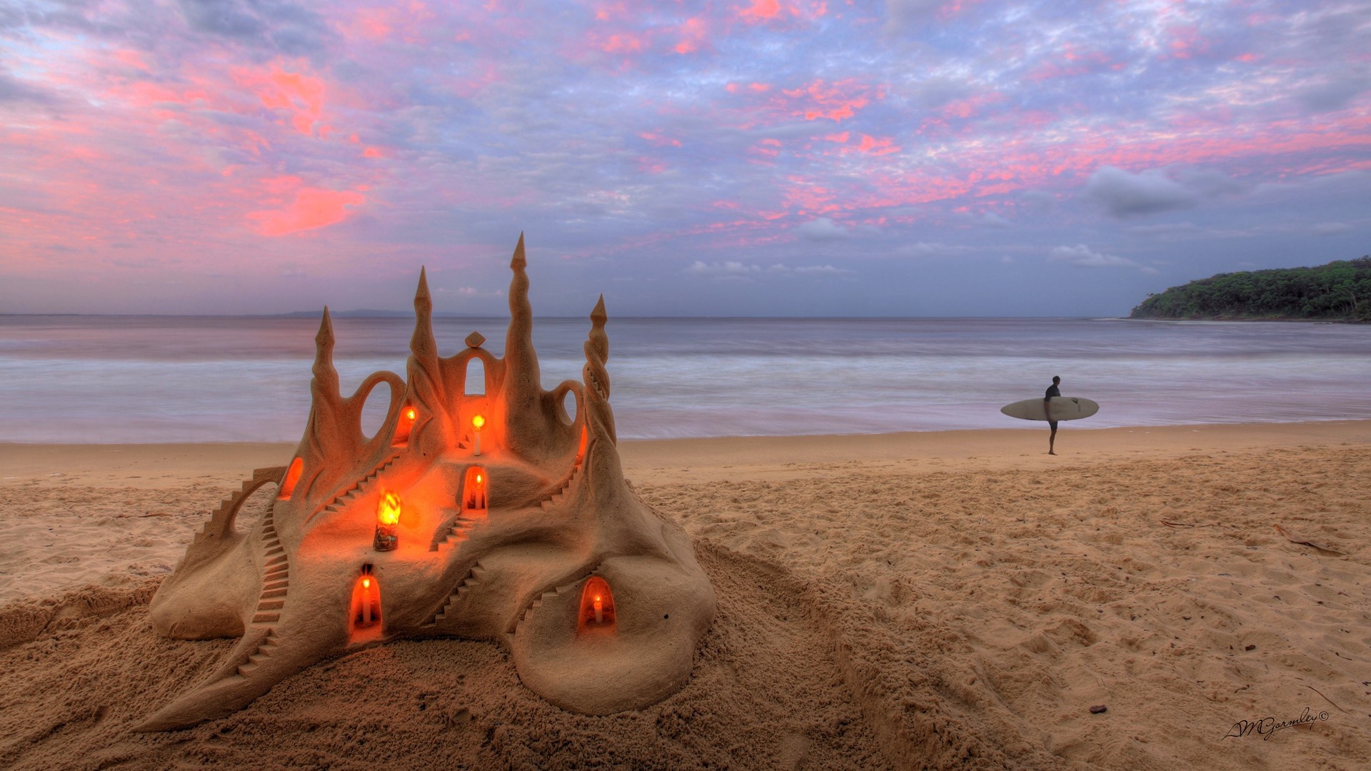 Sandcastle with candles on beach at sunset with surfer HD Wallpaper
