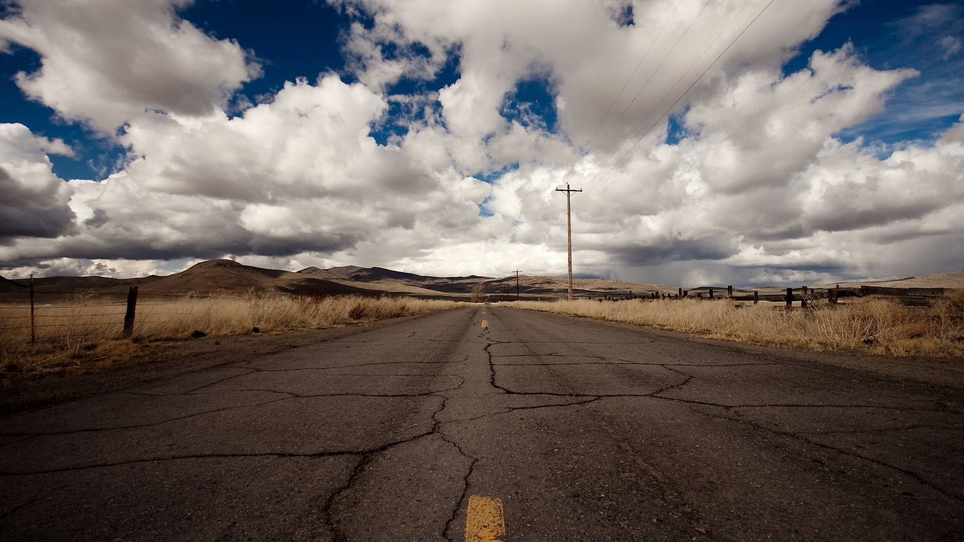 Cracked asphalt road through a dry, grassy landscape under a cloudy sky HD Wallpaper