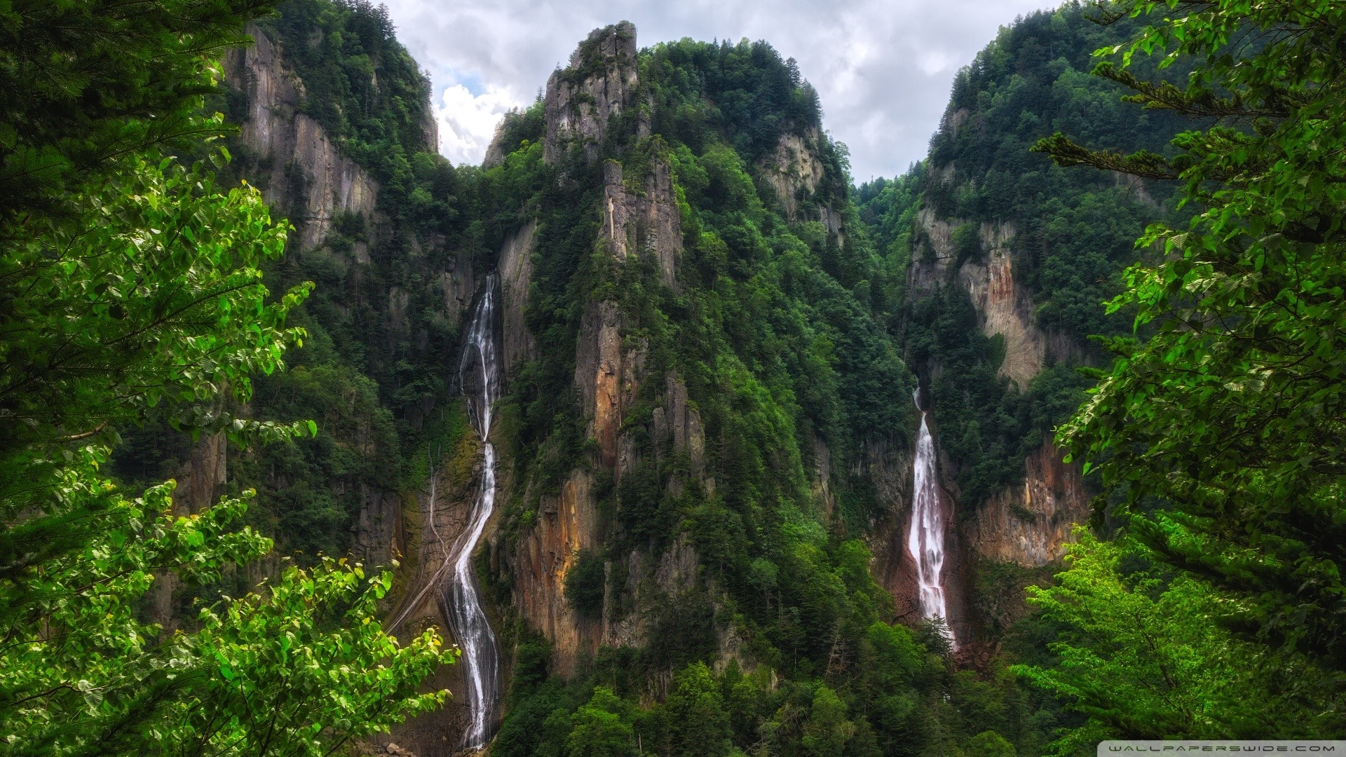 Two waterfalls cascade down a rocky cliff face surrounded by dense green forest. HD Wallpaper