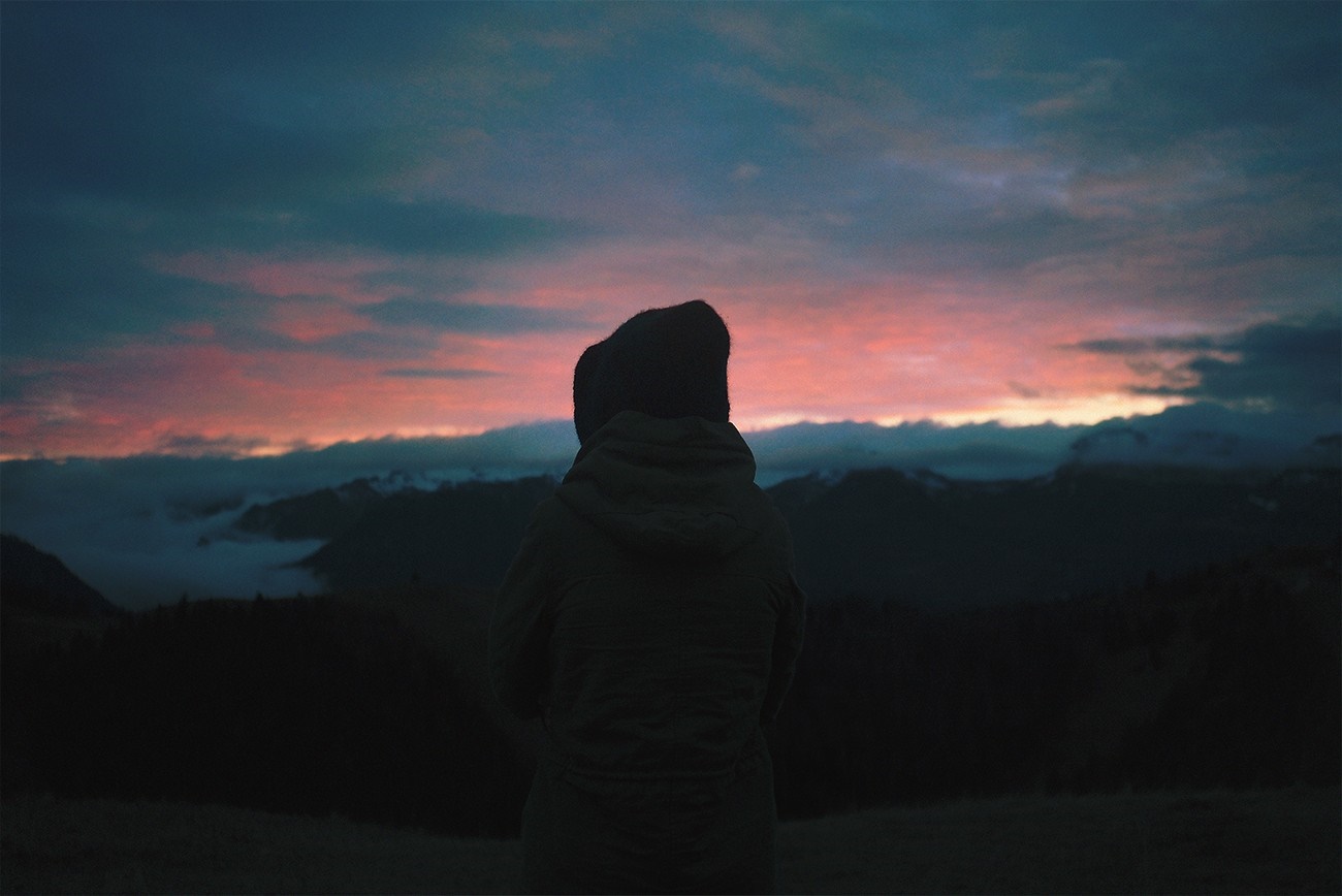 Person in hooded jacket watching sunset over mountains HD Wallpaper
