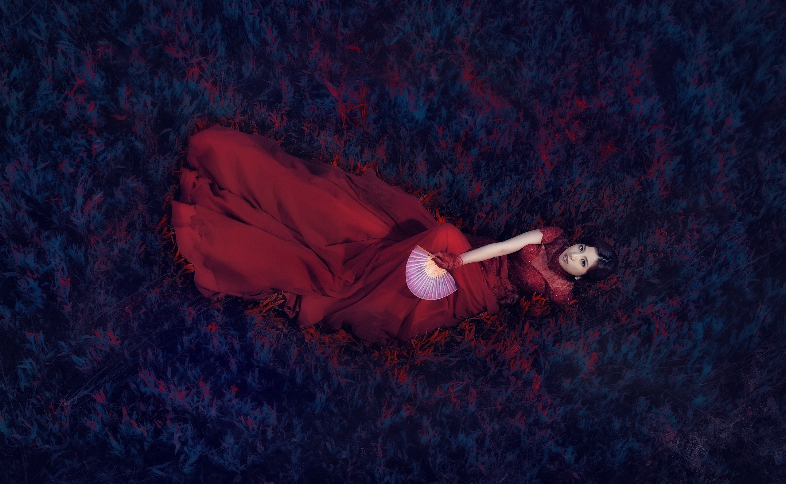 Woman in red dress lying on dark grass holding a fan HD Wallpaper