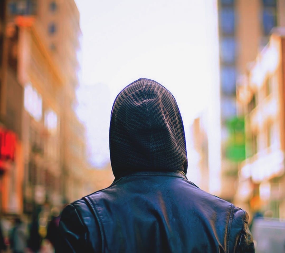 Person wearing a black leather jacket and hood walking down a street HD Wallpaper