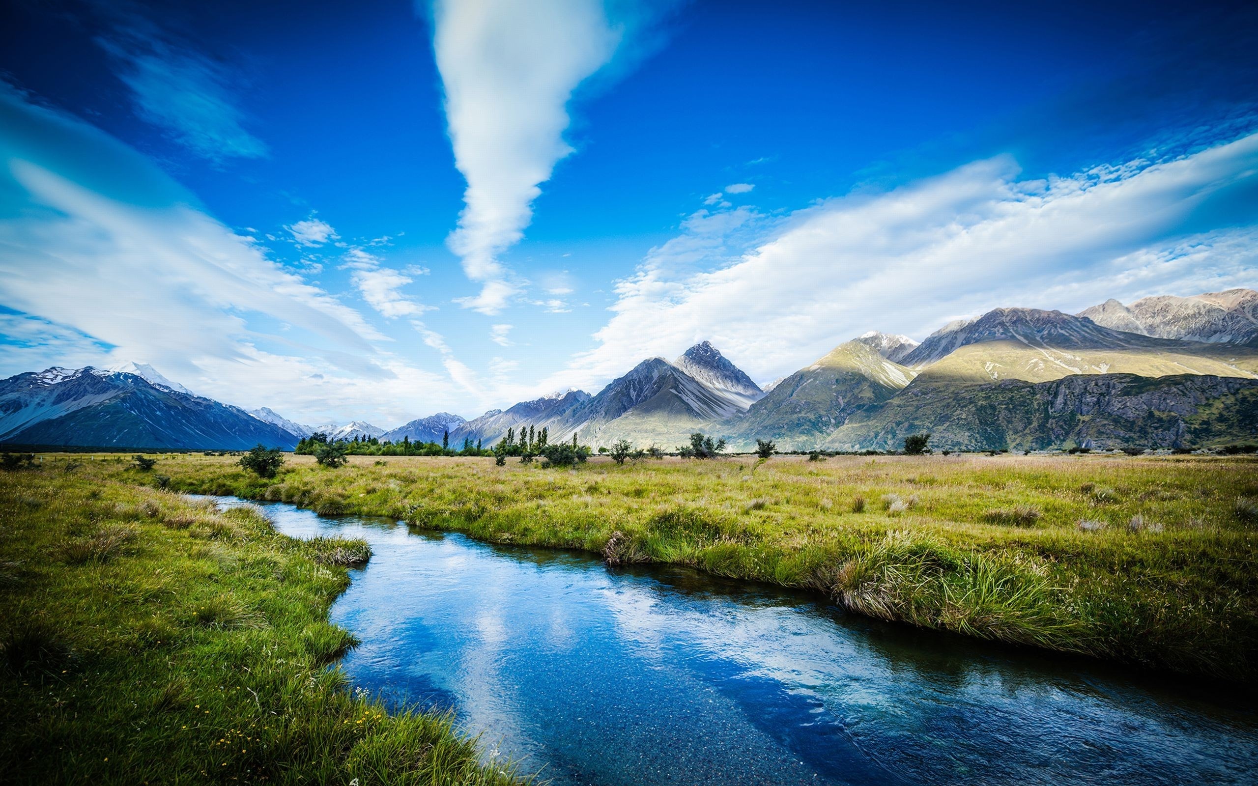 River flowing through a meadow towards mountains under a blue sky HD Wallpaper