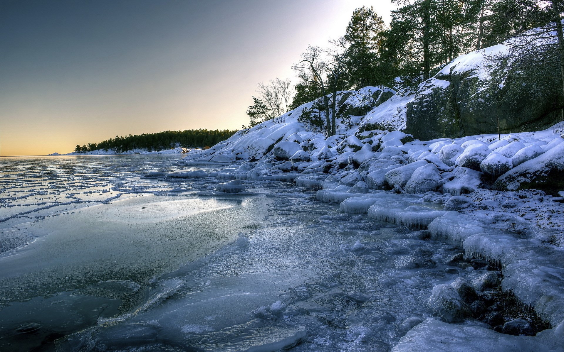 Frozen lake shore with snow-covered rocks and trees HD Wallpaper